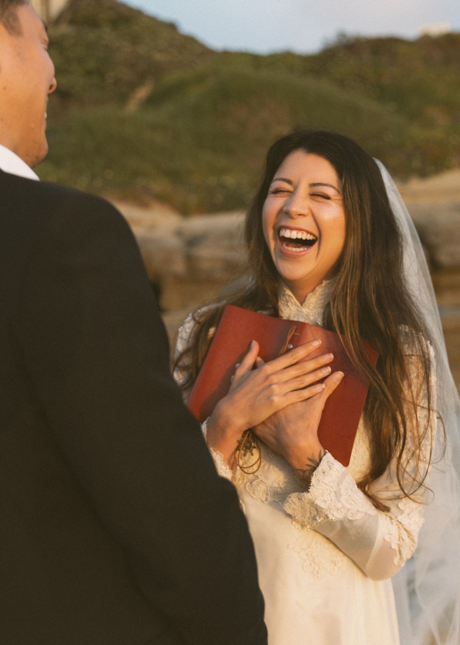 Bride in lace wedding dress and veil laughing joyfully during outdoor beach wedding ceremony.