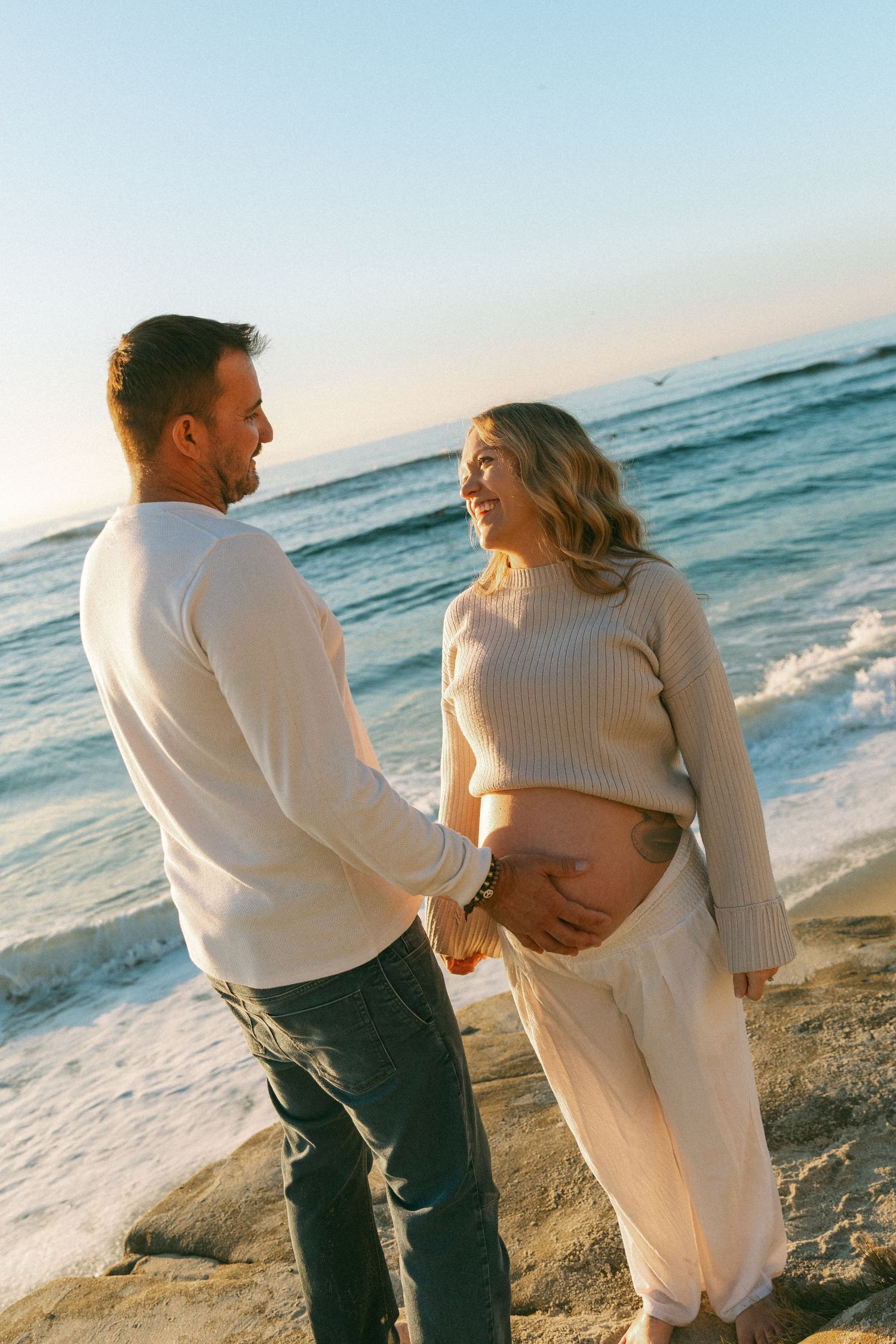 Maternity photoshoot on beach with couple in white sweaters against ocean backdrop.
