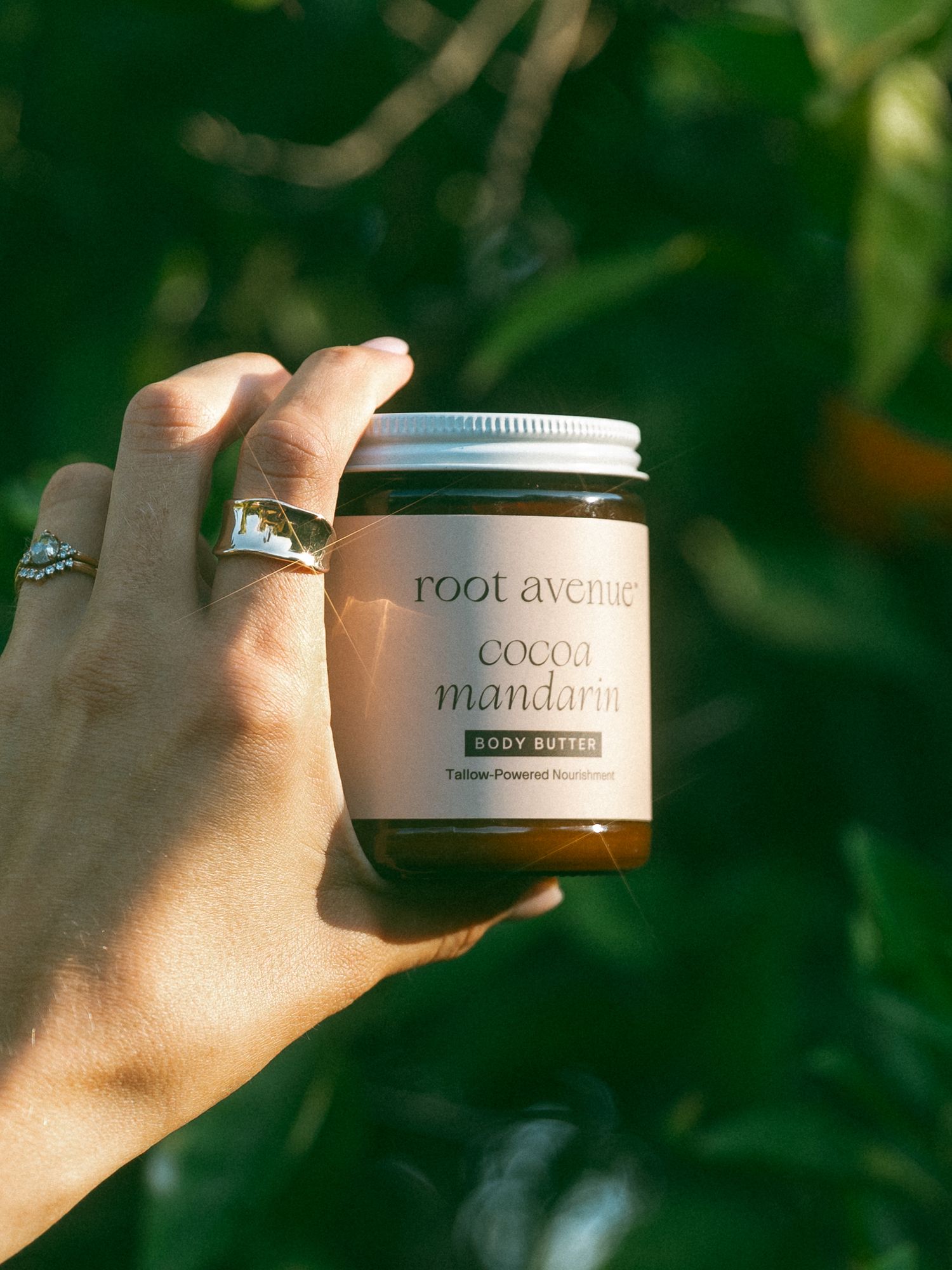Hand holding a jar of cocoa mandarin body butter with rings visible against a blurred green background.