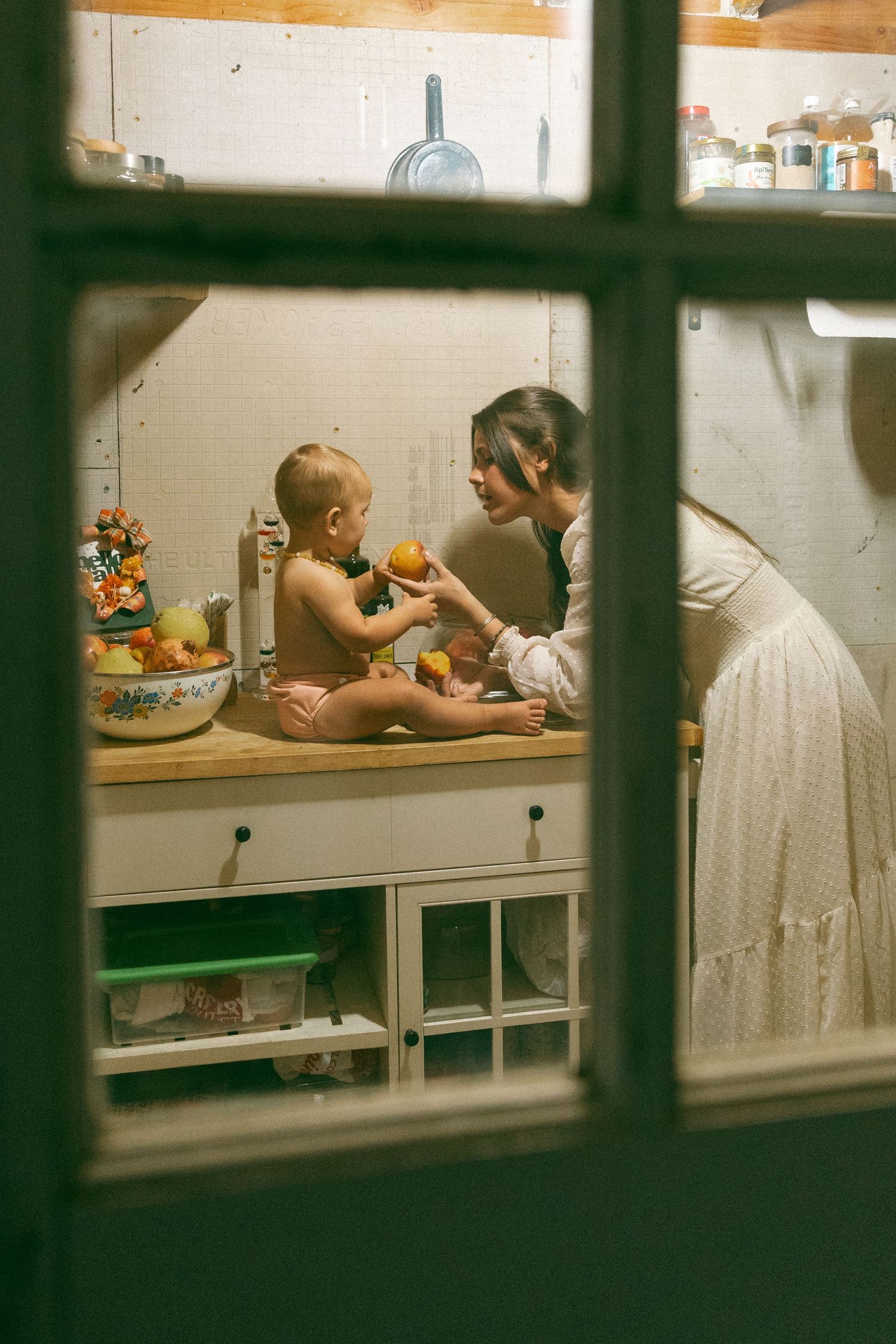 Baby lying on a changing table with toys and storage shelves visible through window panes.