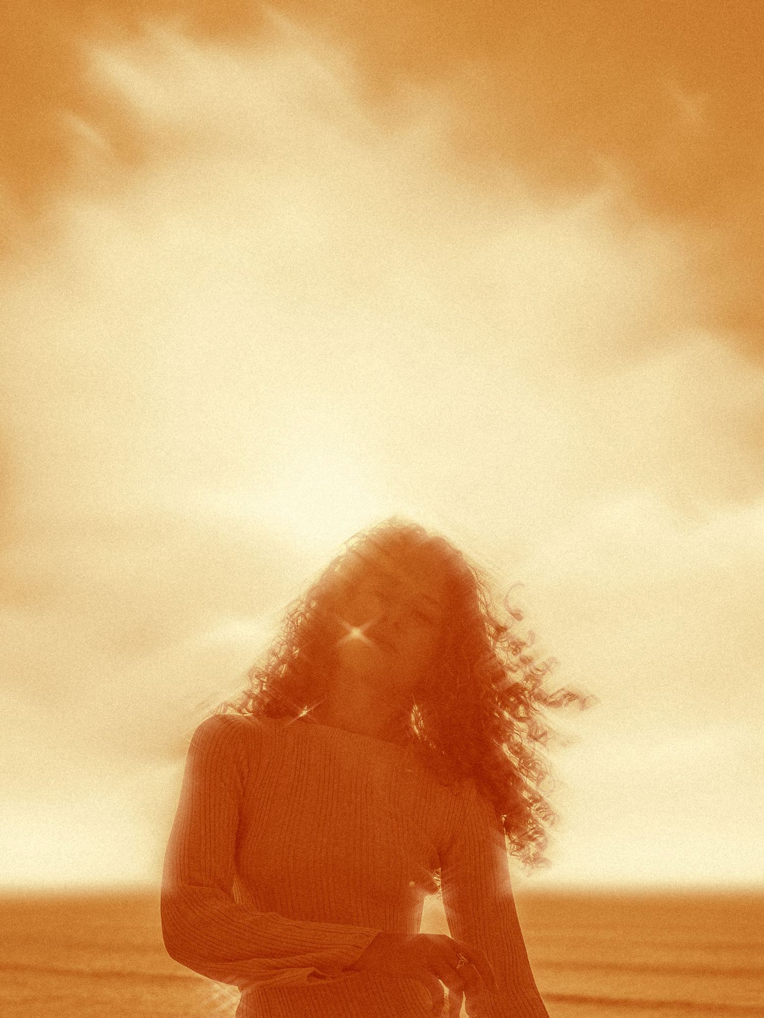 Dreamy sepia portrait with curly hair backlit by golden sunset sky over ocean.