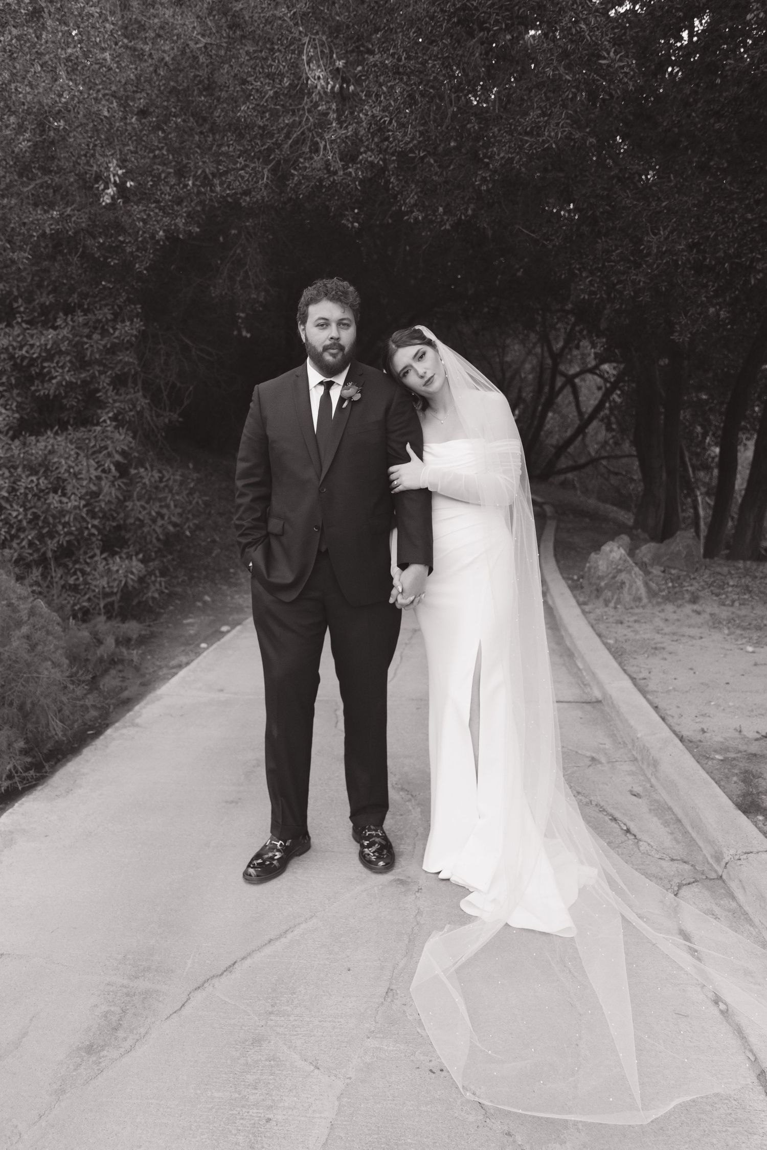 Black and white wedding photo on a path surrounded by trees.