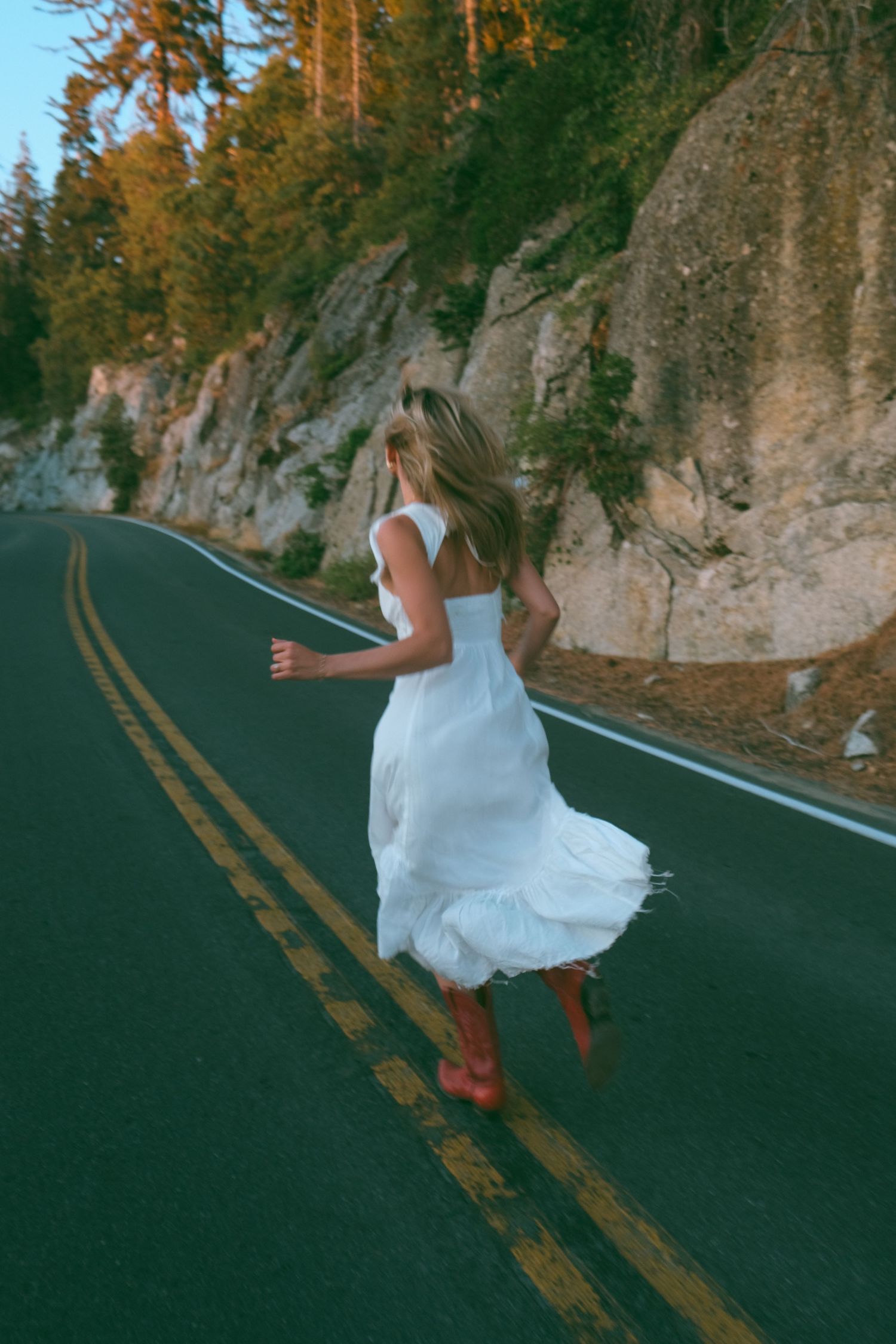 Figure in flowing white dress runs along a winding mountain road at sunset.