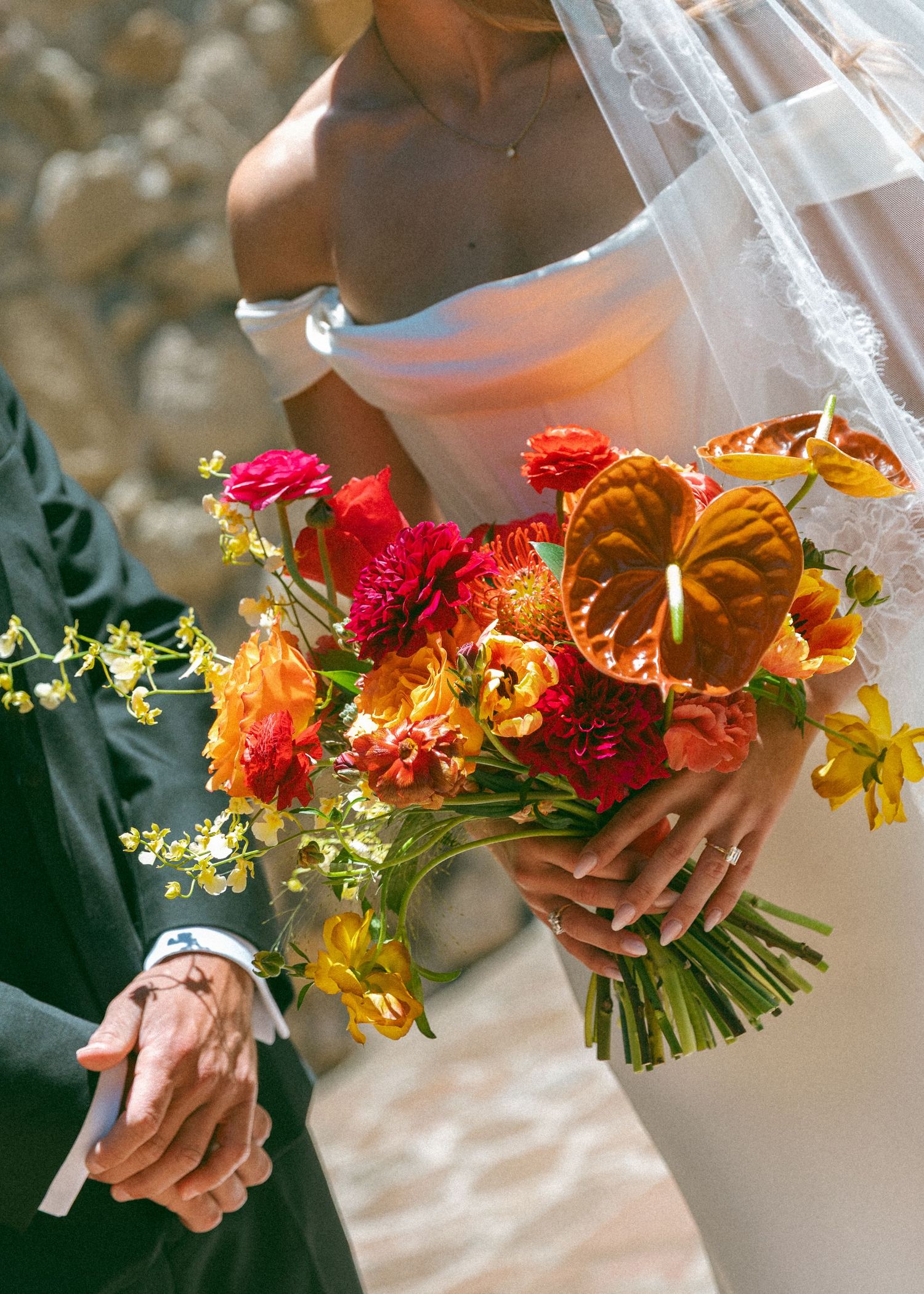 Bride holding vibrant orange, red and yellow wedding bouquet with anthurium flowers while groom holds her hand.