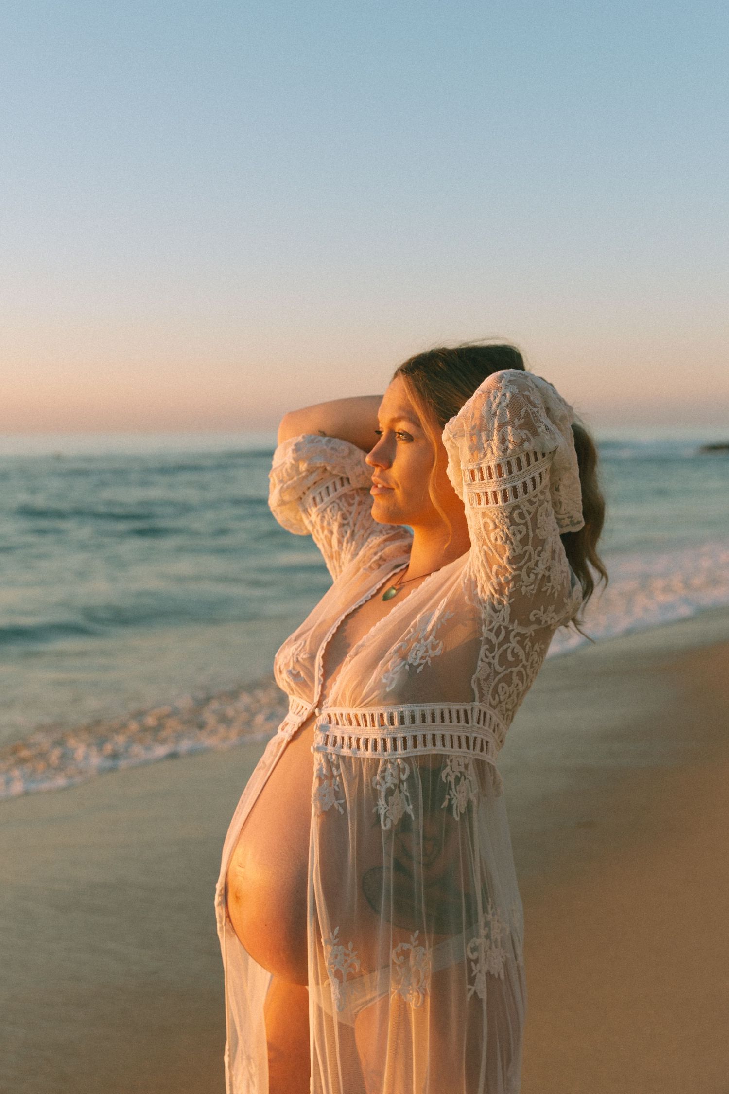 Ethereal maternity pose in sheer lace dress on sandy beach at sunset.