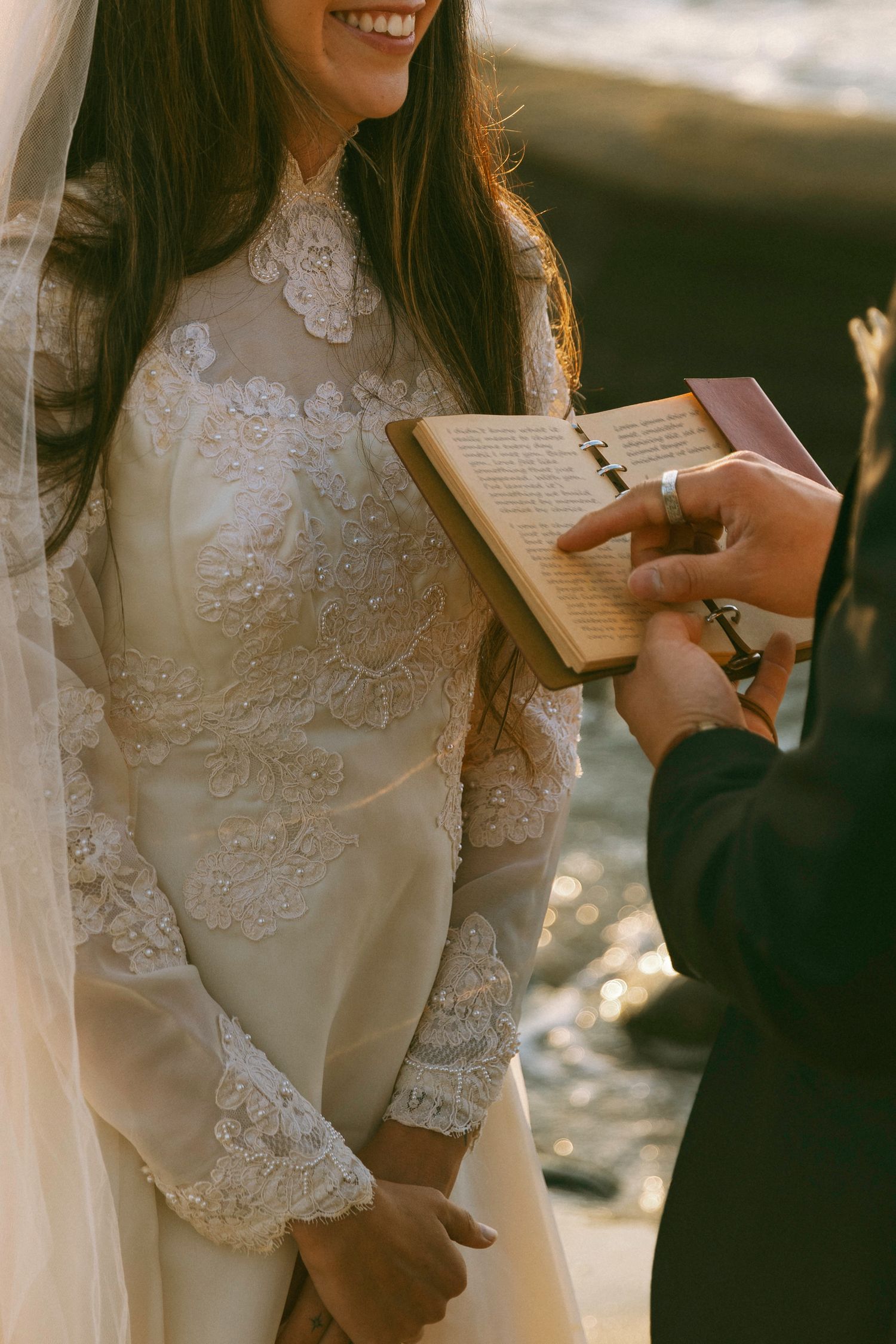 Smiling bride in lace wedding dress holding vows booklet during outdoor ceremony.