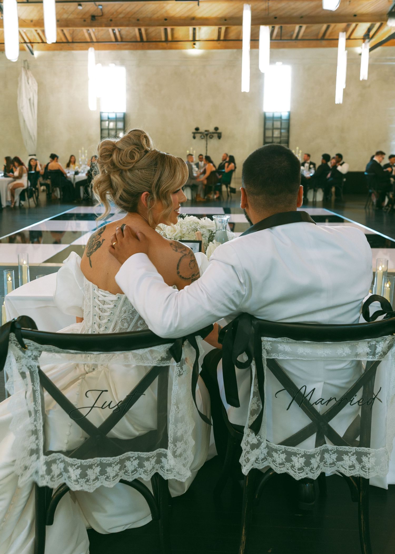 Bride and groom seated at sweetheart table viewed from behind, chairs decorated with lace and ribbon.