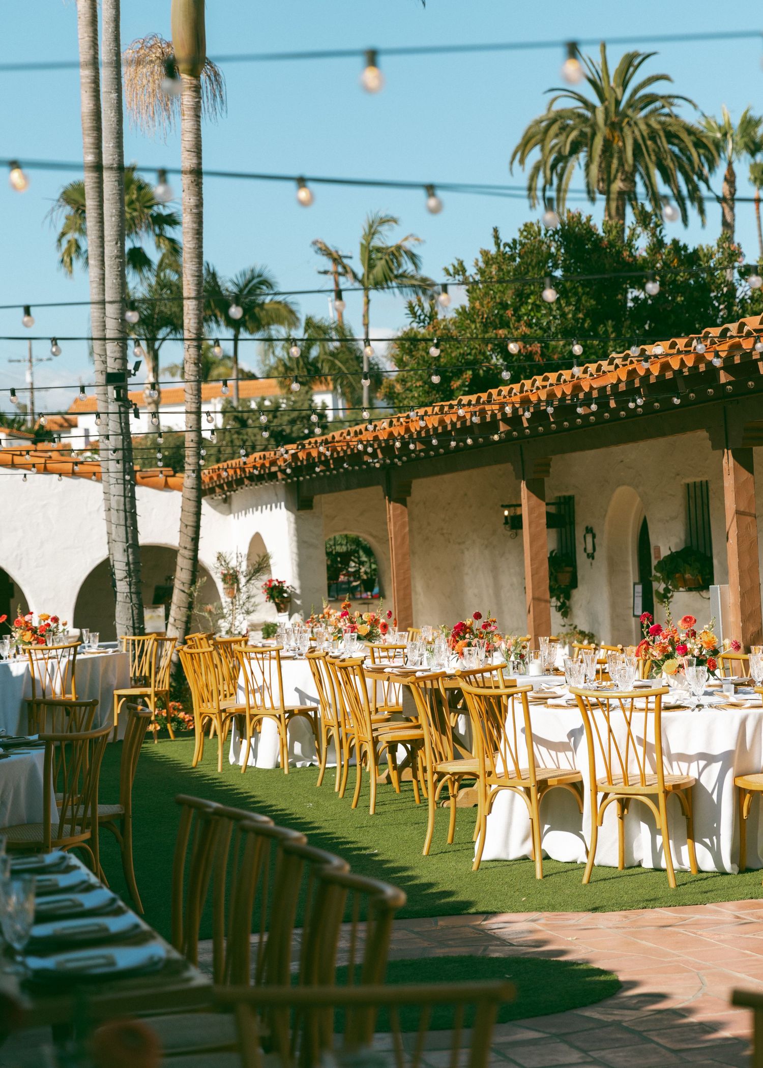 Outdoor wedding reception with long tables, gold cross-back chairs, string lights and palm trees at Spanish venue.