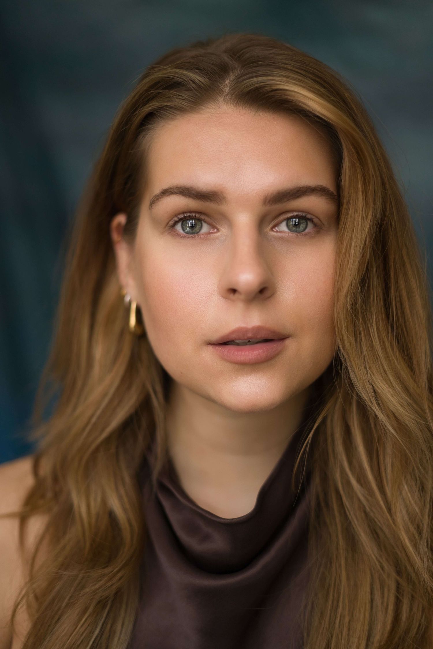 Portrait photo of someone with long honey blonde hair and blue eyes wearing a brown turtleneck against a dark background.