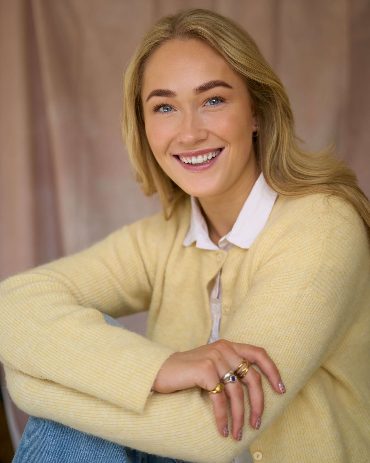 Person wearing a pastel yellow sweater and white collar smiles warmly while leaning on their arm.