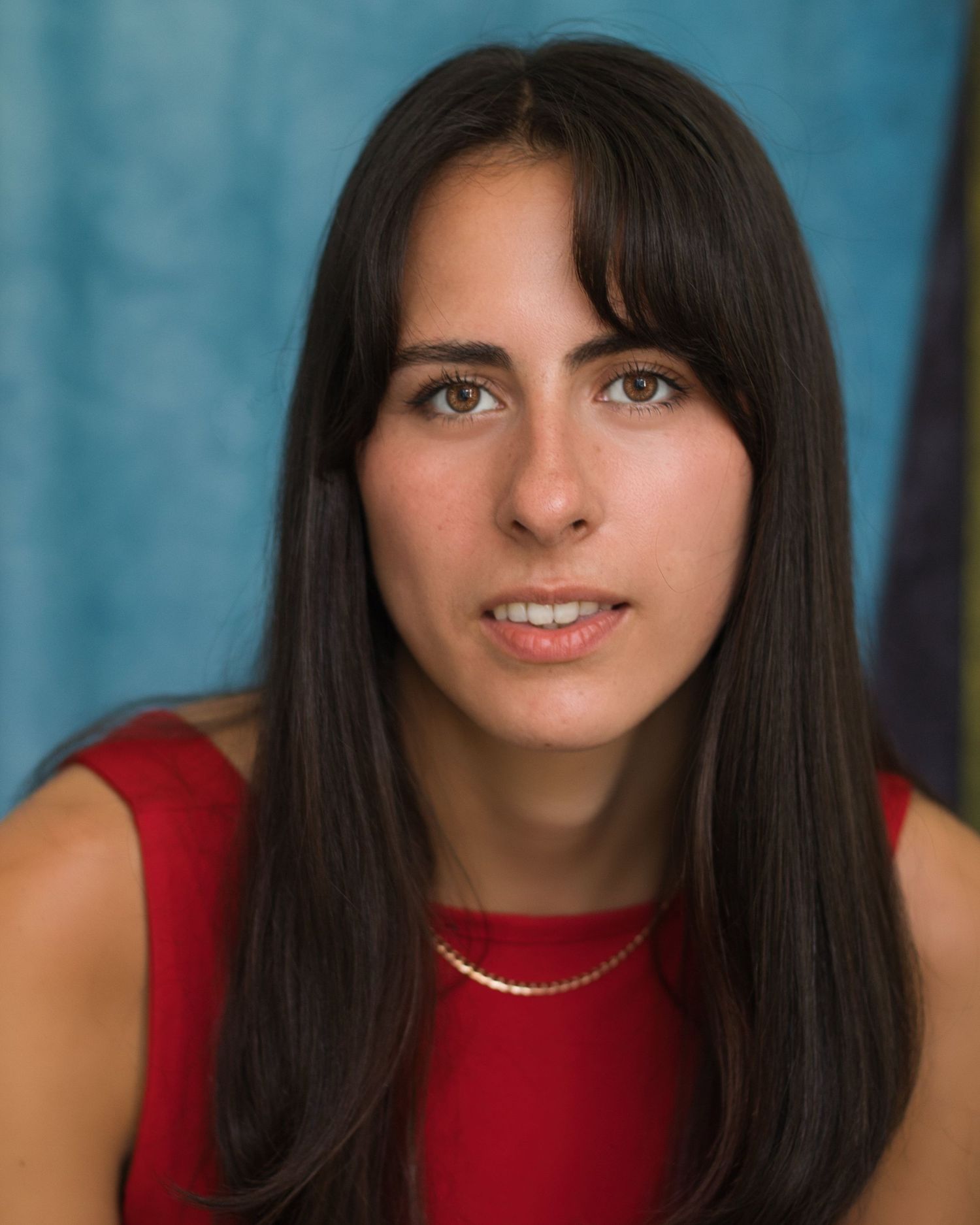 A woman in a red sleeveless top smiles warmly at the camera in a professional headshot series against a blue background.