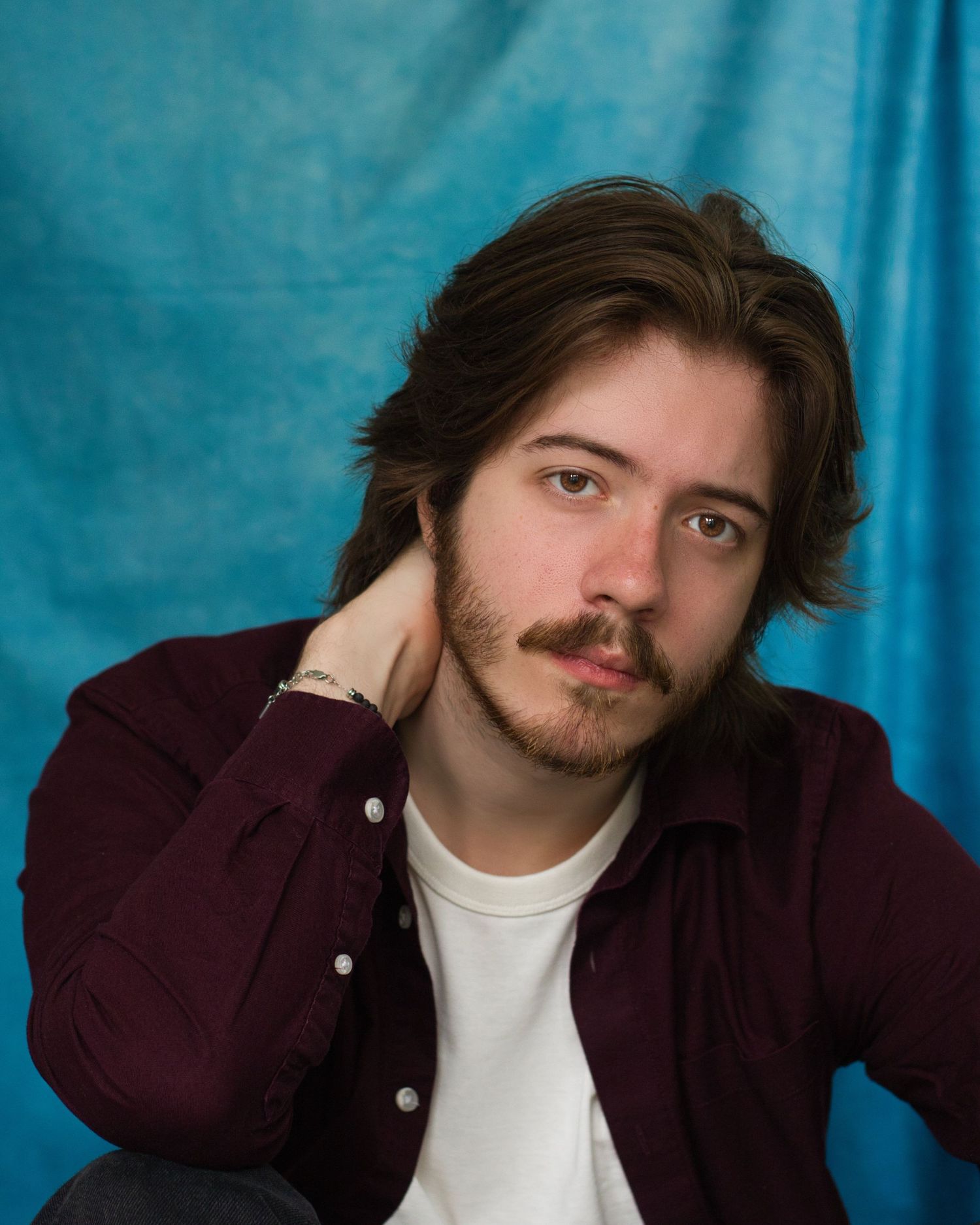 Portrait against a turquoise backdrop showing someone in a burgundy button-up shirt and white t-shirt with dark hair and facial hair.