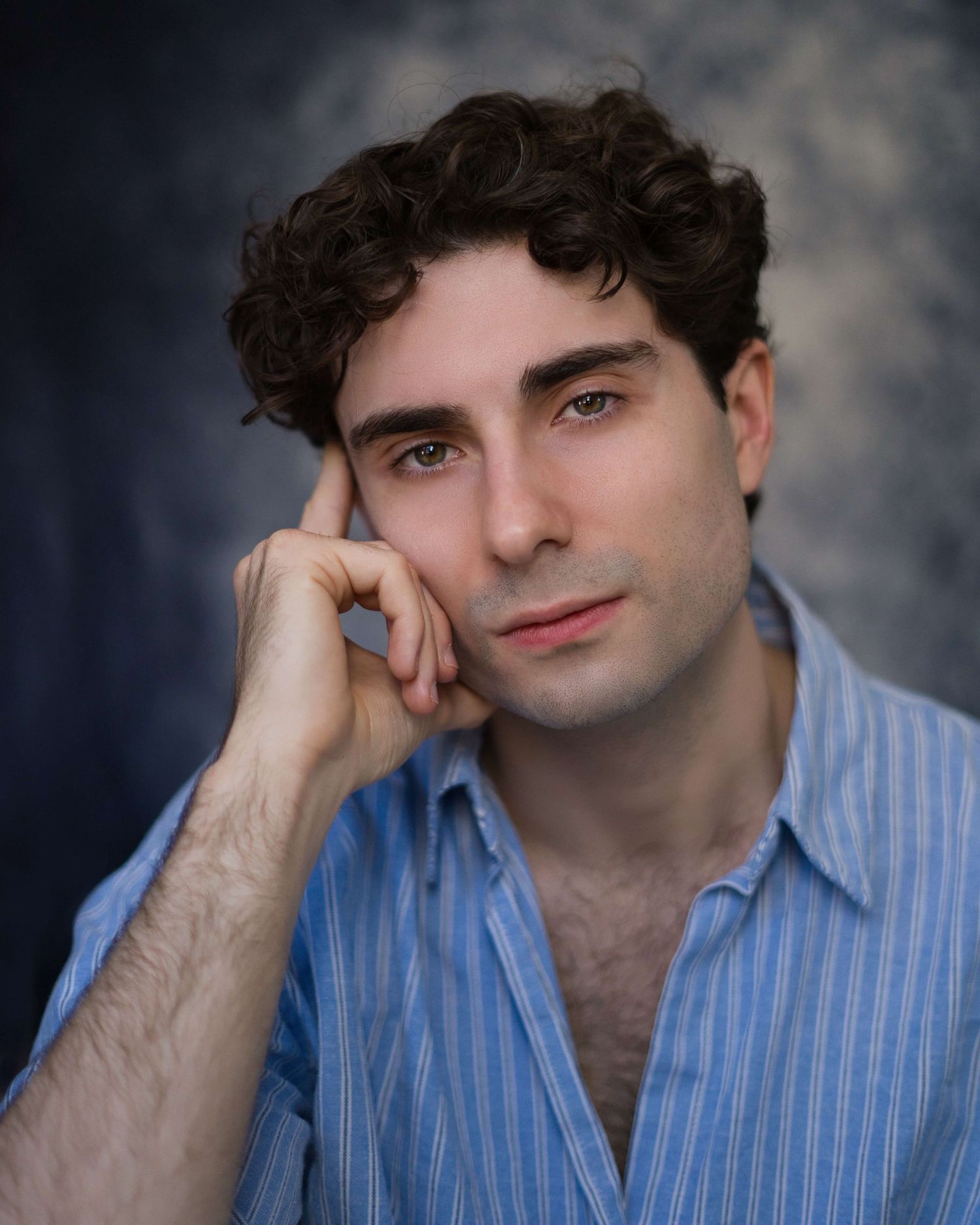 A person in a blue striped shirt poses against a dark background in a professional headshot series.