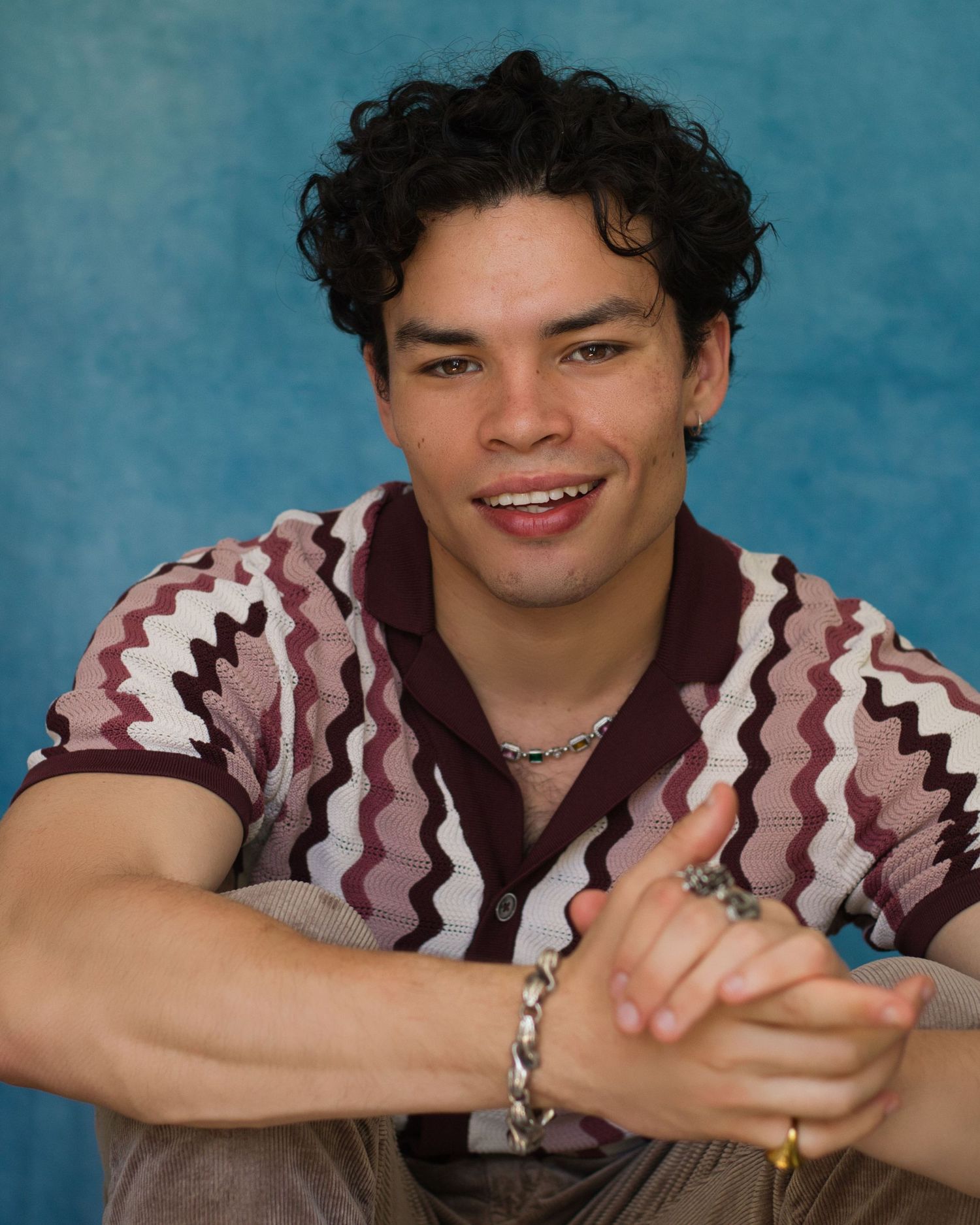 A person wearing a brown and white zigzag patterned shirt smiles while posing against a blue background.