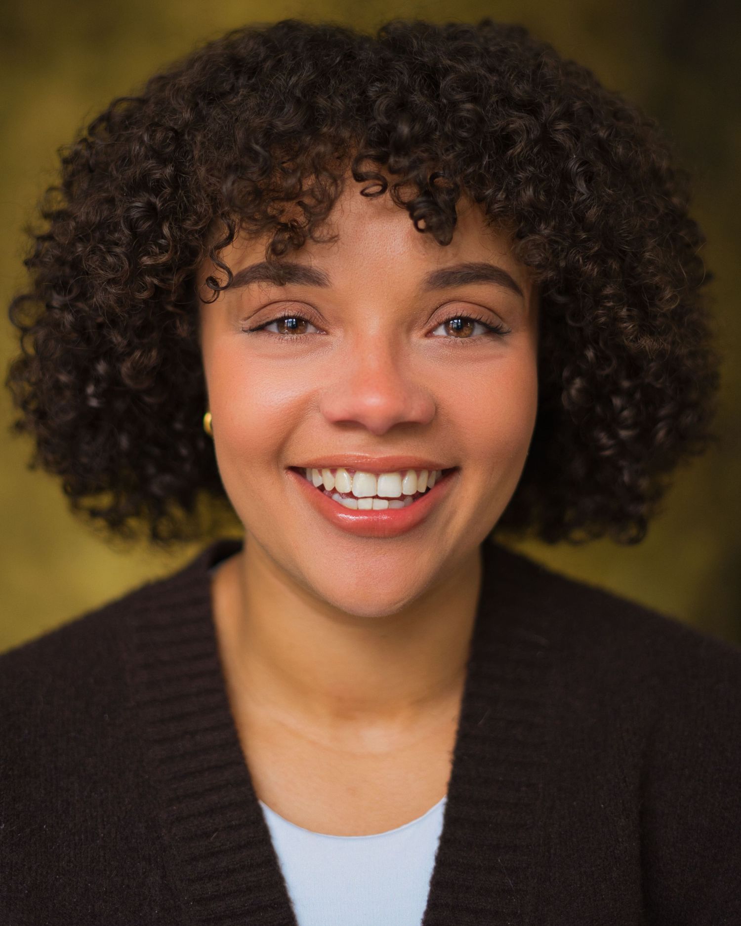 Young woman with curly hair smiling brightly, wearing a dark cardigan against a green background.