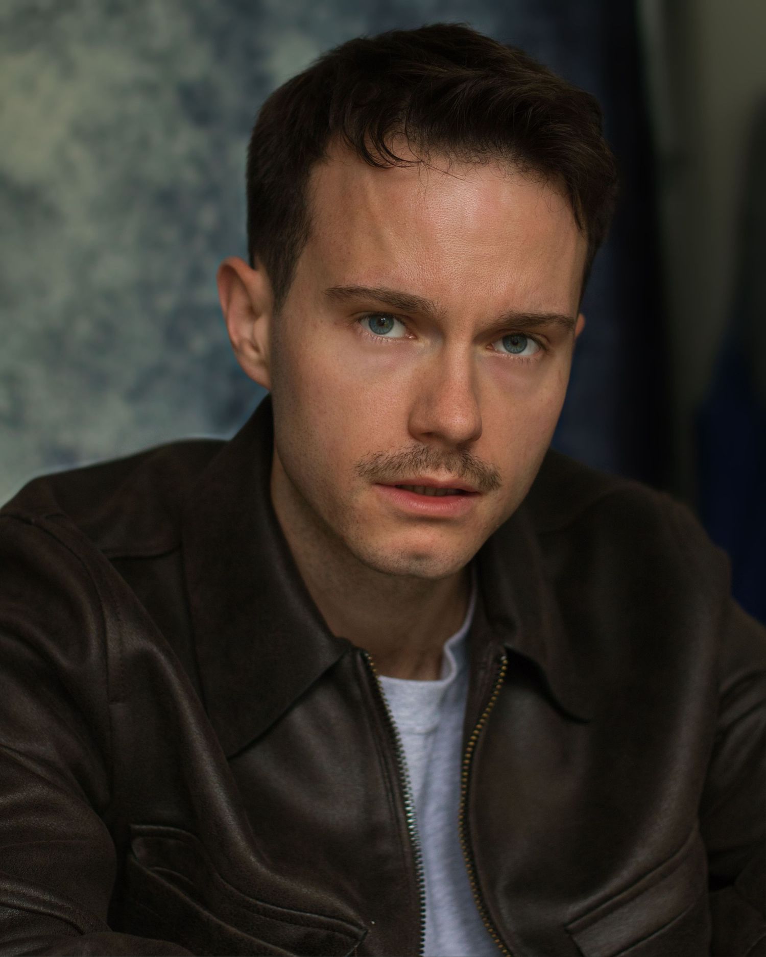A person wearing a brown leather jacket and white t-shirt gazes intently with dramatic lighting in a dark setting.