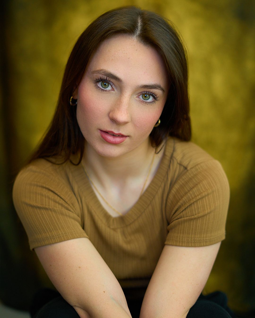 Young woman with green eyes and brown hair poses in a tan ribbed top against a golden bokeh background.