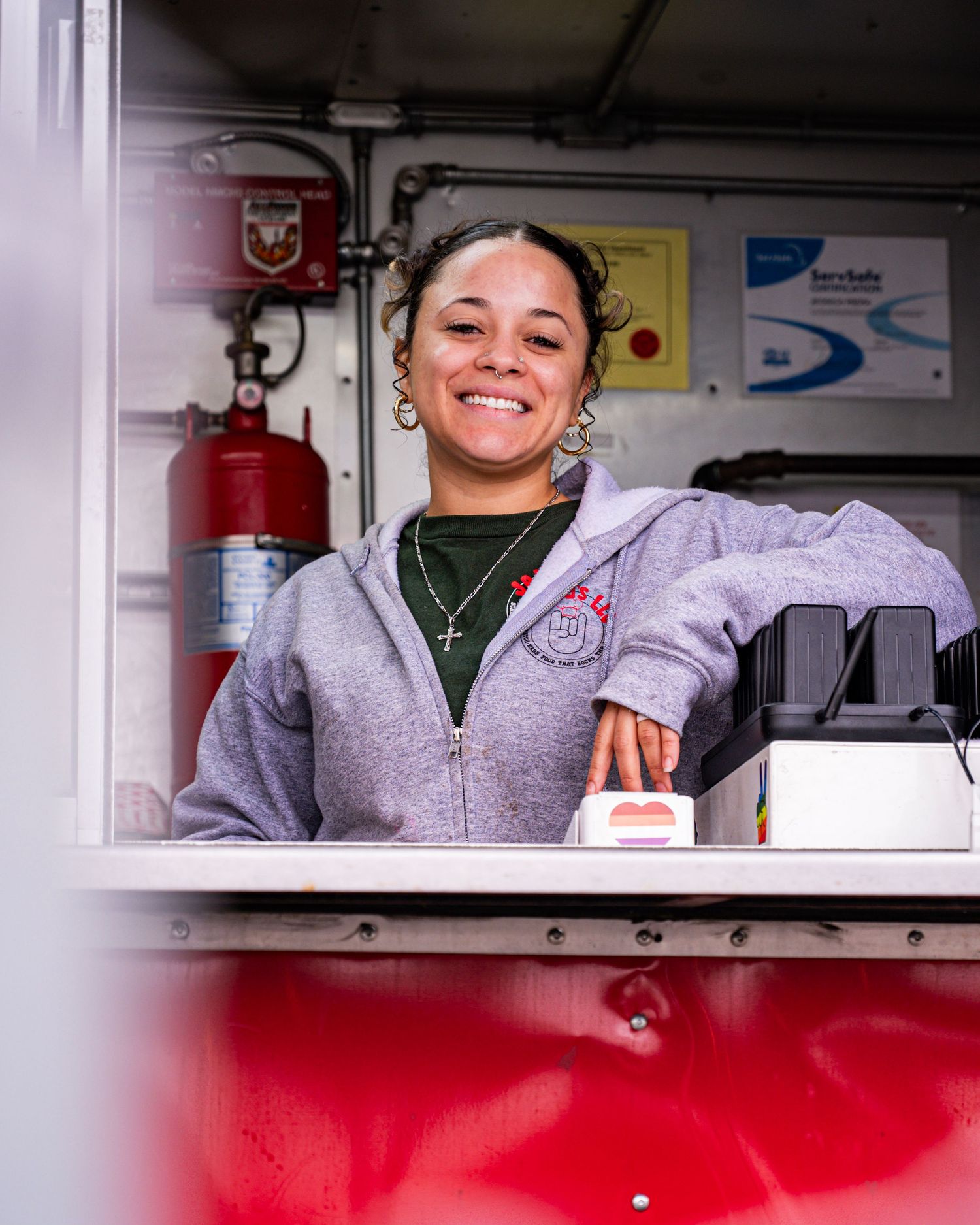 Smiling woman in gray hoodie working inside a red food truck with fire suppression equipment visible.