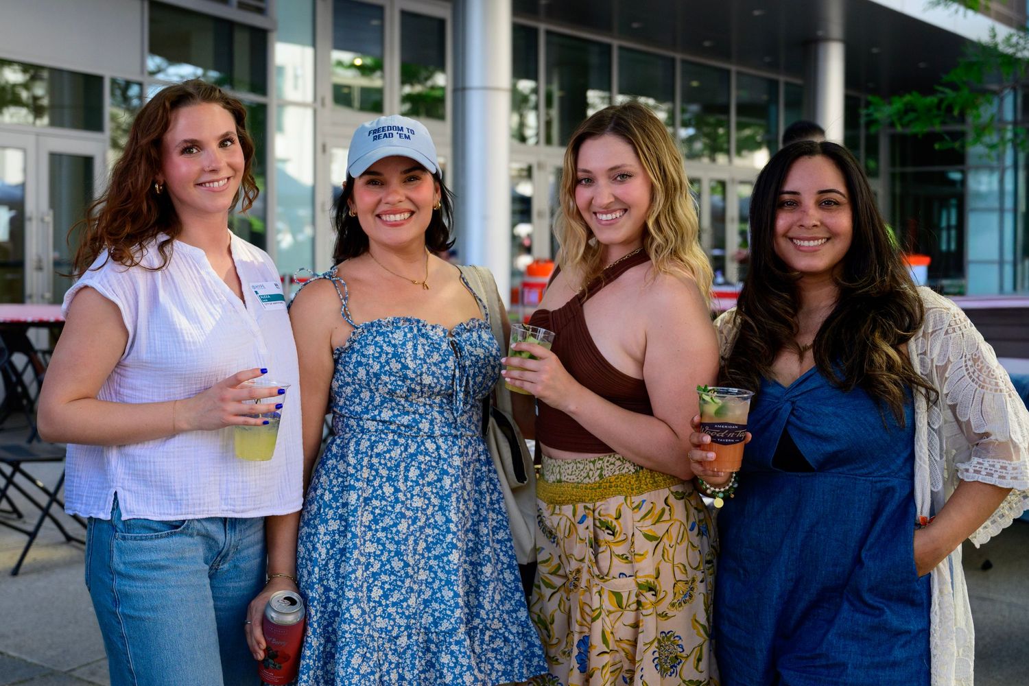 Four women smiling and holding drinks at an outdoor social event on a sunny day.