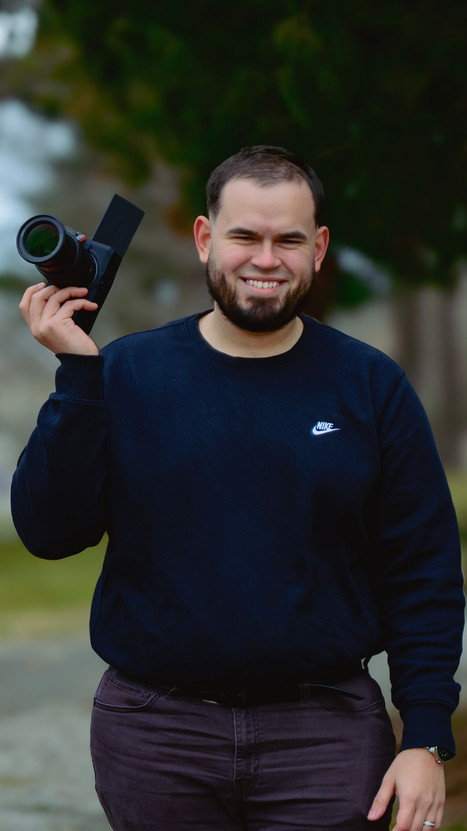 Smiling man holding a camera, wearing a navy Nike sweatshirt, standing outdoors with blurred green trees in background.