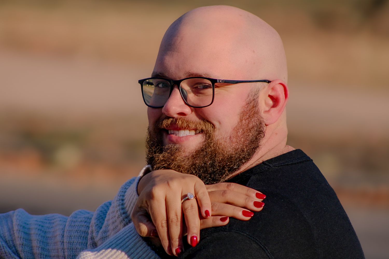 A person with glasses and a beard wearing black, resting their chin on their hand against a blurred outdoor background.