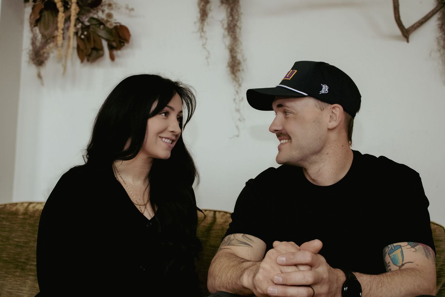 Two people in black clothing sit close together and share a candid moment while smiling at each other.