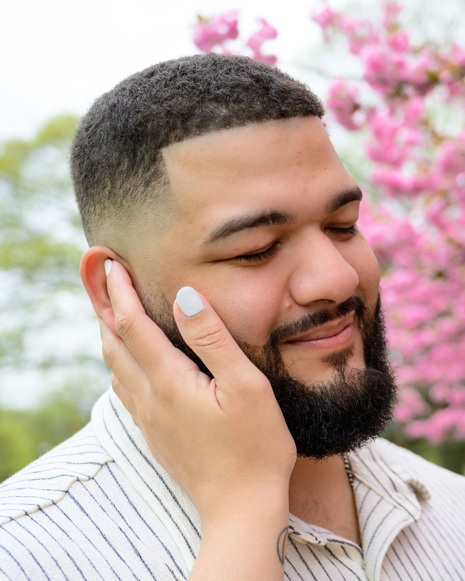 A bearded man with a fade haircut smiles softly, hand on cheek, with pink cherry blossoms in the background.