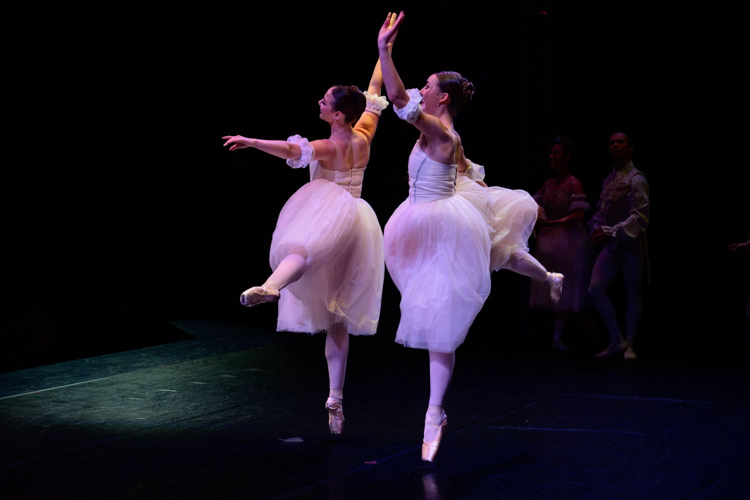 Two ballerinas in white tutus perform an elegant dance pose on stage under dramatic lighting.