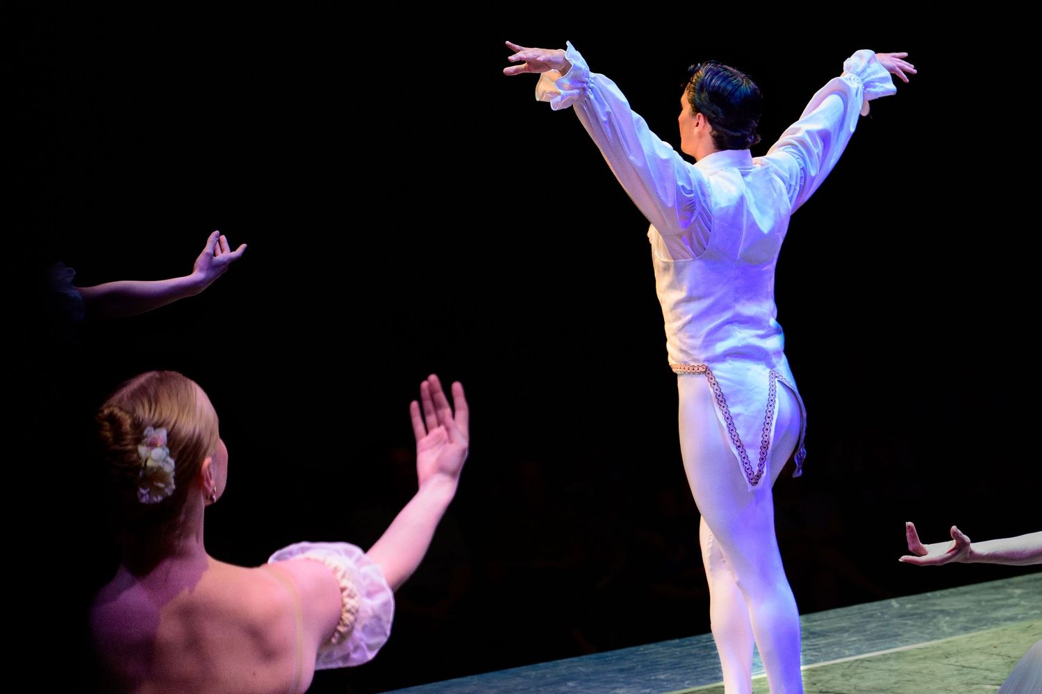 Two ballet dancers perform on stage, one in white costume with arms raised gracefully under dramatic lighting.
