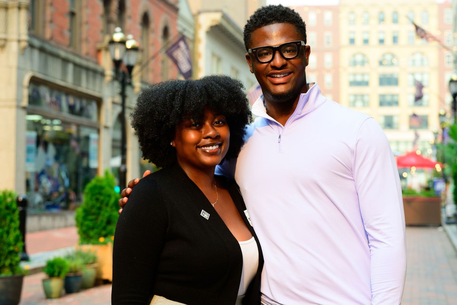 A smiling couple poses together on a city street, woman in black blazer and man in pink shirt wearing glasses.