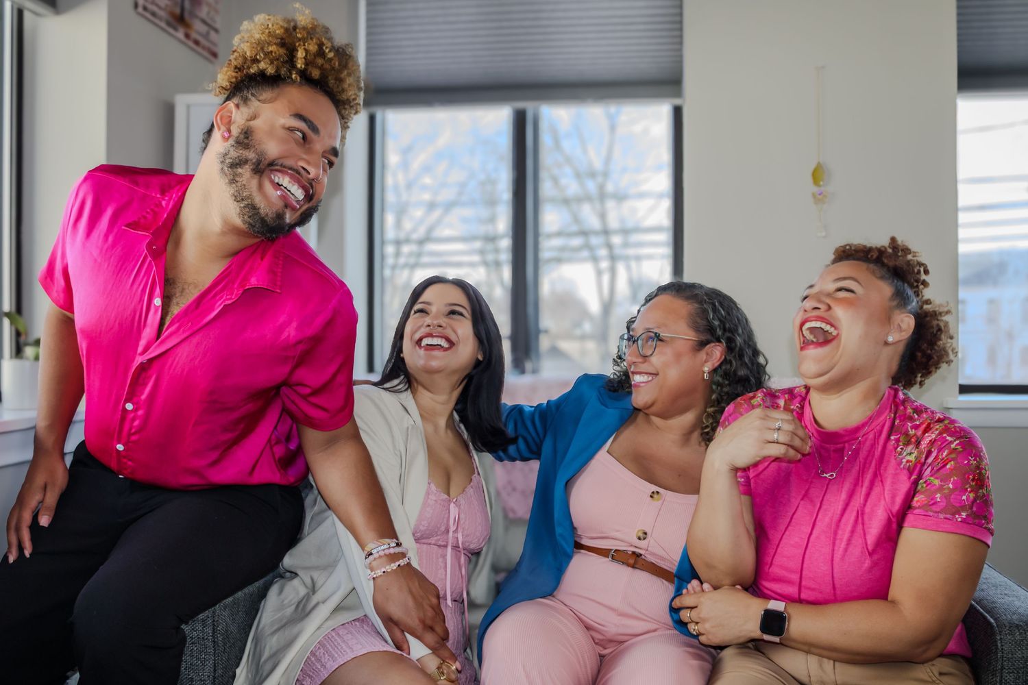 A group of friends laugh together while sitting on a couch, wearing various shades of pink clothing.