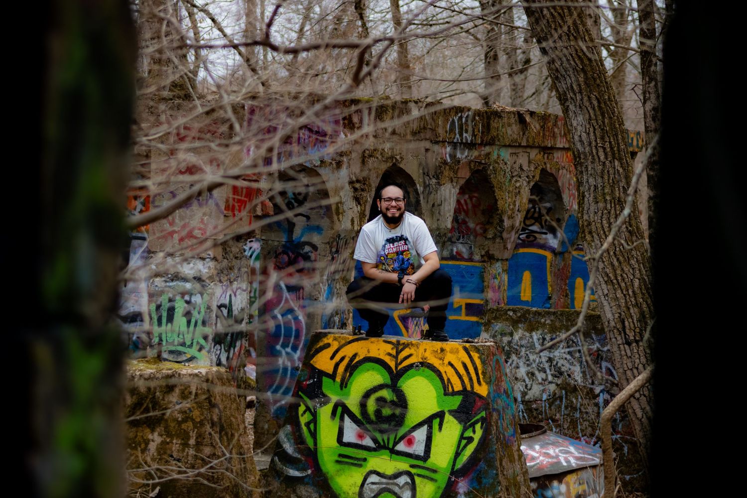Graffiti art featuring a neon green character decorates a concrete wall in a wooded area with person sitting above.