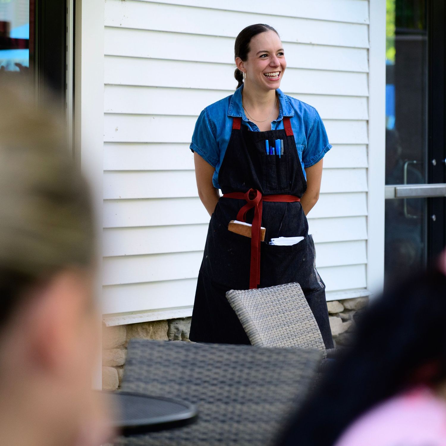 Smiling waitress in black apron and blue shirt standing outdoors at a restaurant patio.