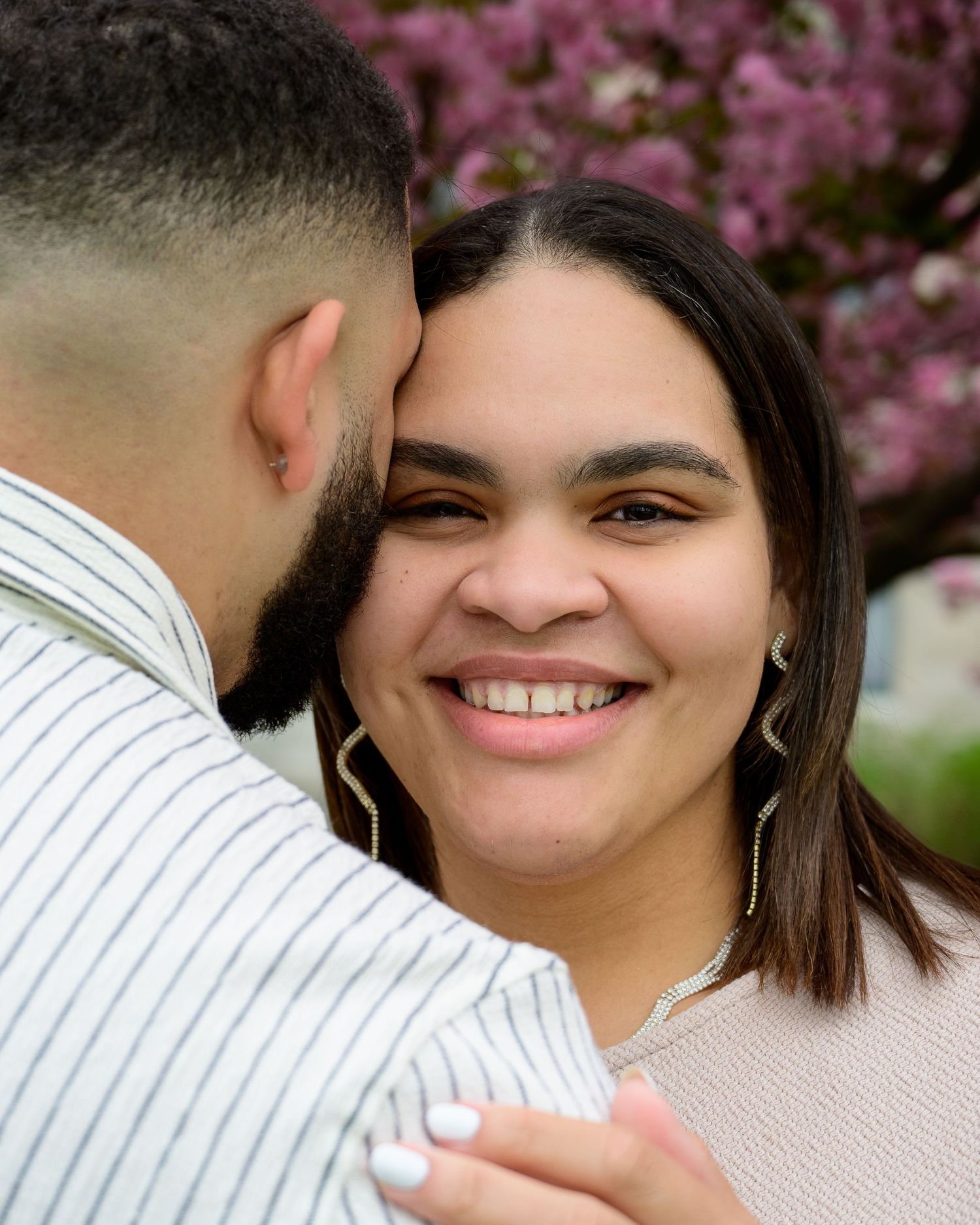 A smiling woman with gold earrings embraces a man outdoors near blooming pink cherry blossom trees in spring.