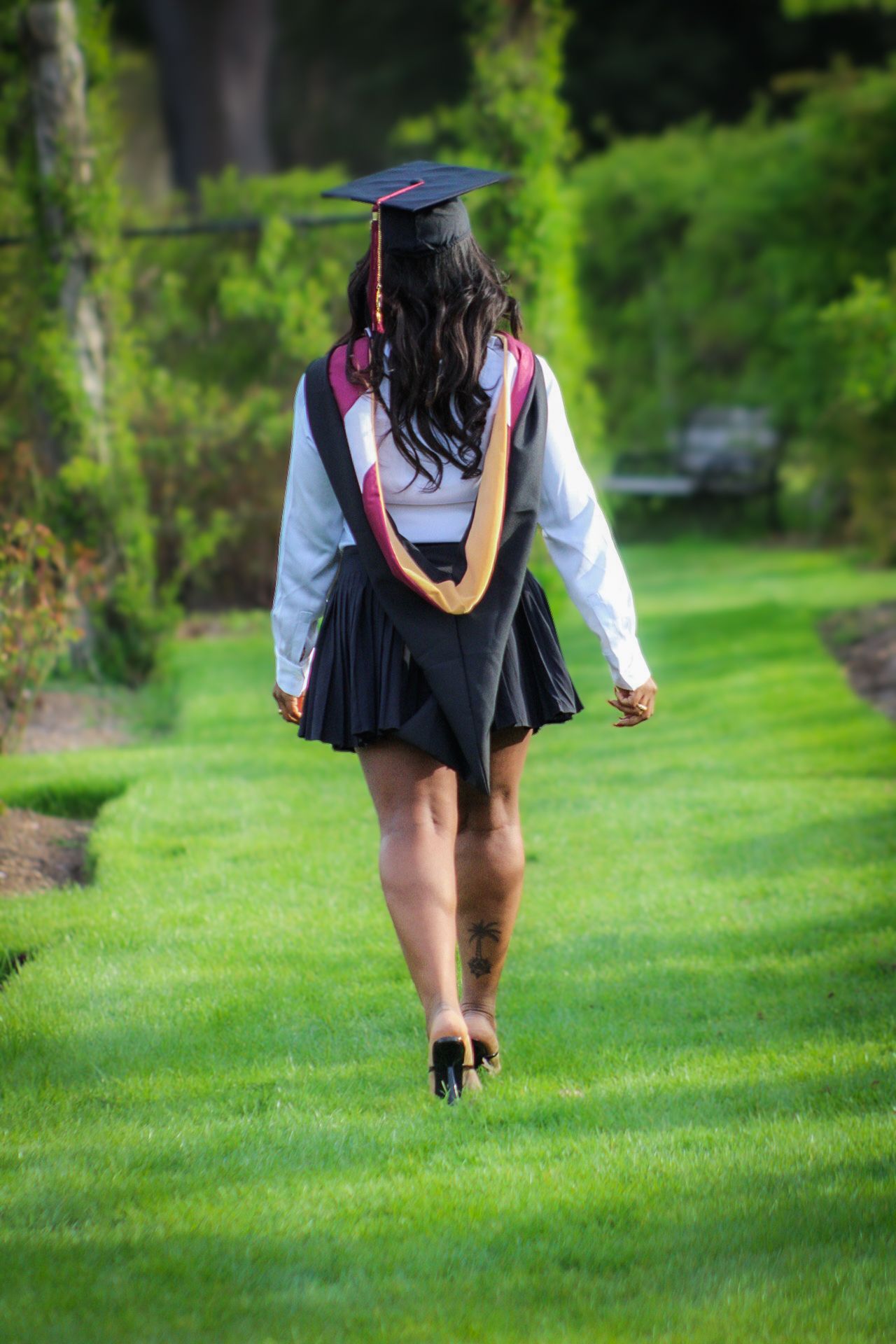 Graduate in cap and gown walking away on a lush green lawn surrounded by trees and gardens.