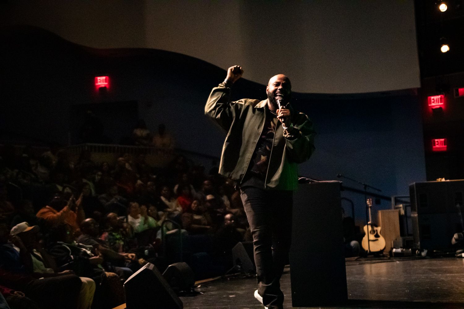 Speaker performs on dimly lit stage with red exit signs visible in background.