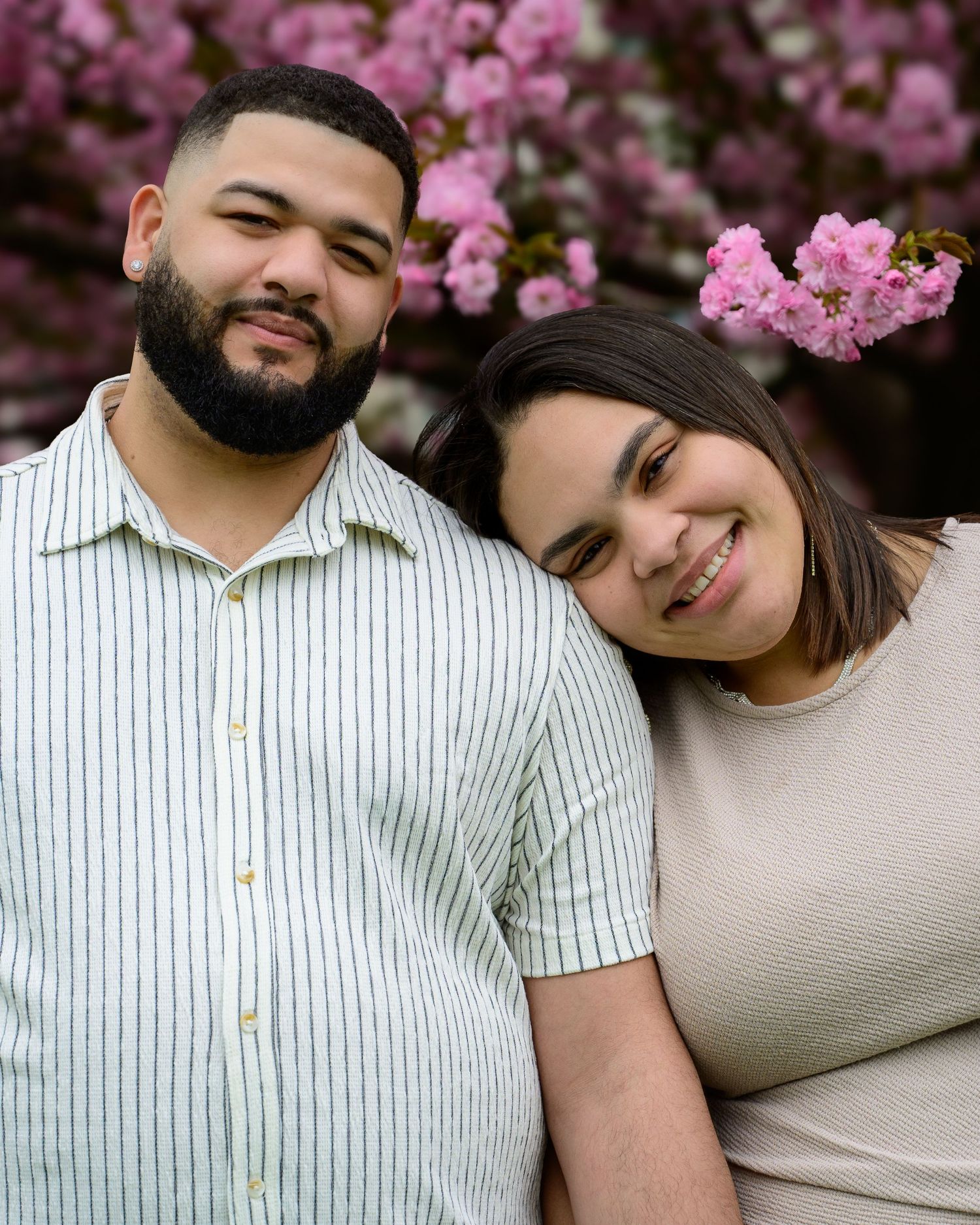 A couple smiles together in front of pink cherry blossom trees, the man with a beard wearing a striped shirt.