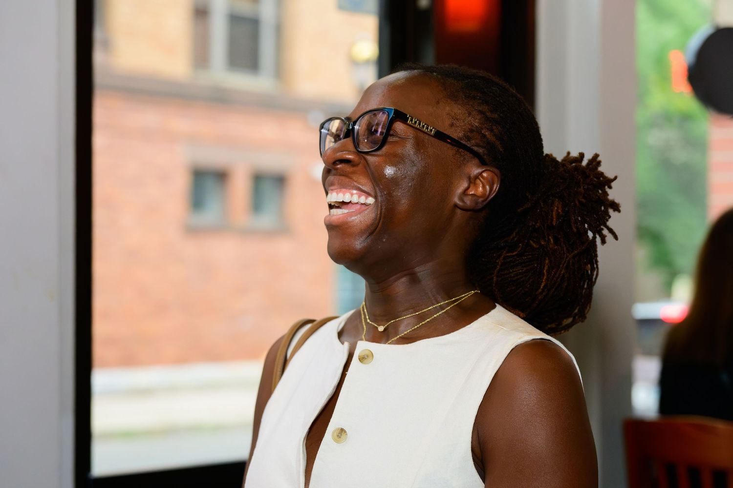 A joyful person with dreadlocks and glasses laughing in a bright indoor setting near a window.
