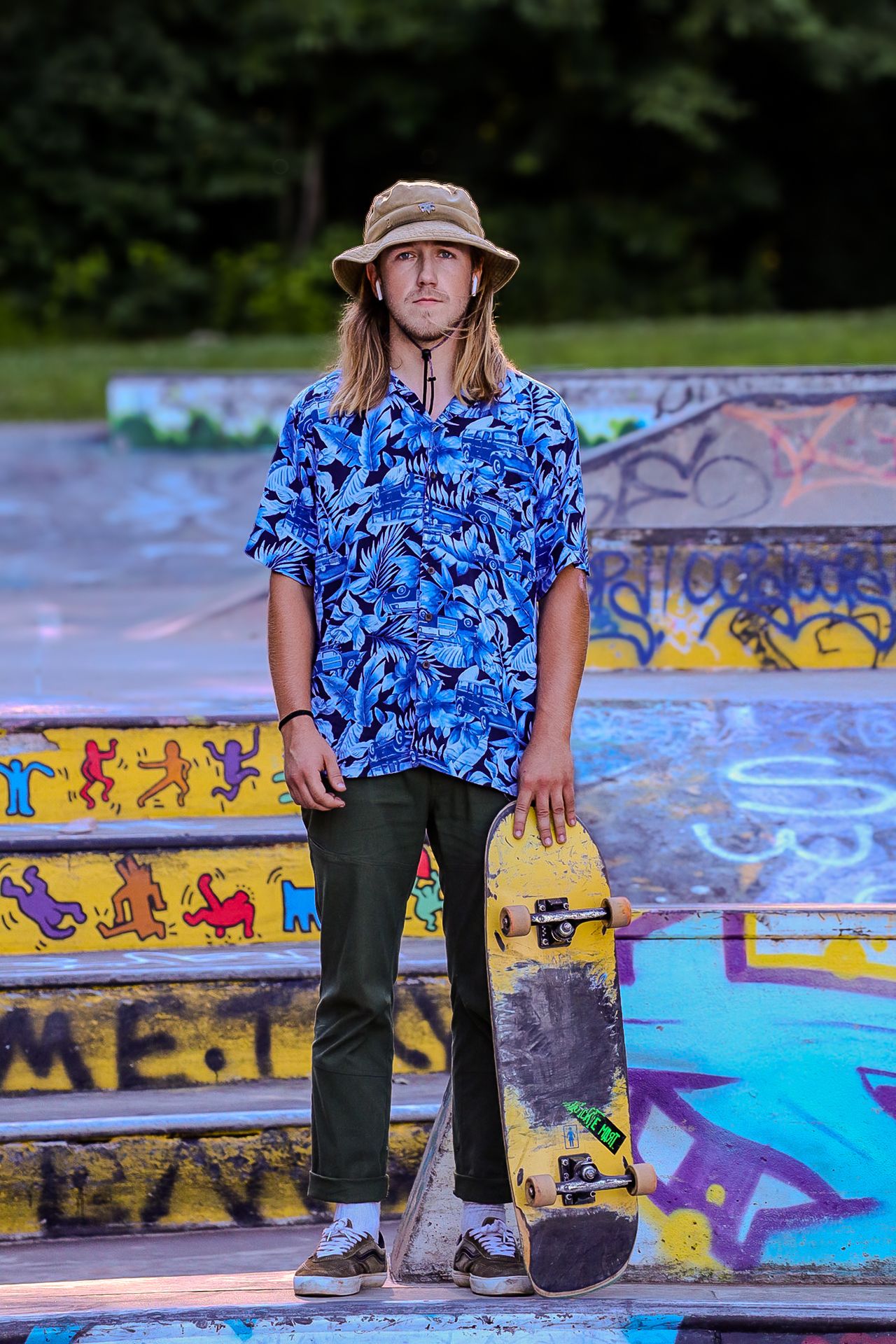 Skateboarder in blue patterned shirt and bucket hat standing at colorful skate park with graffiti.
