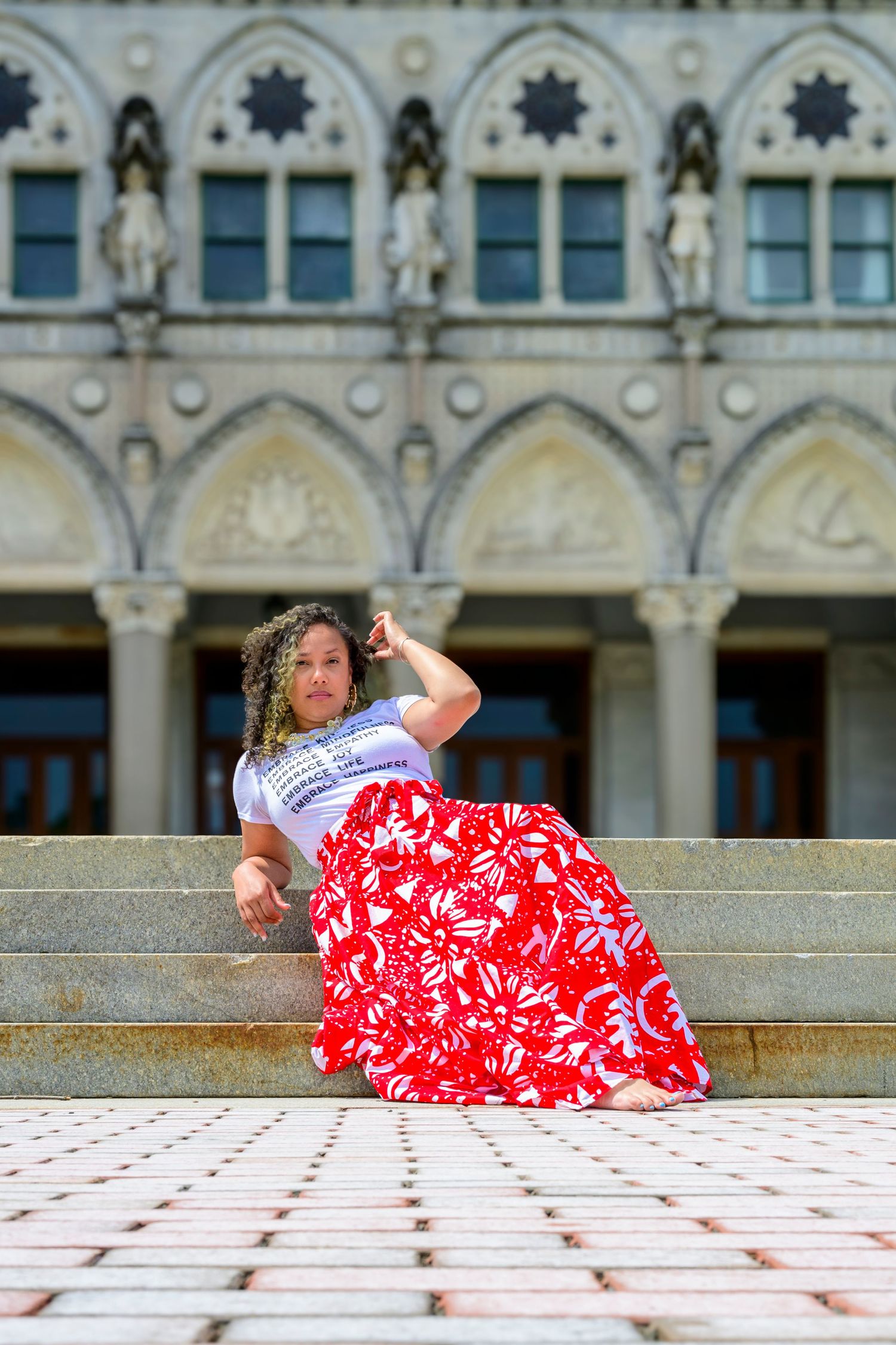 Person in red and white floral skirt lounging on stone steps of ornate Gothic architecture building.