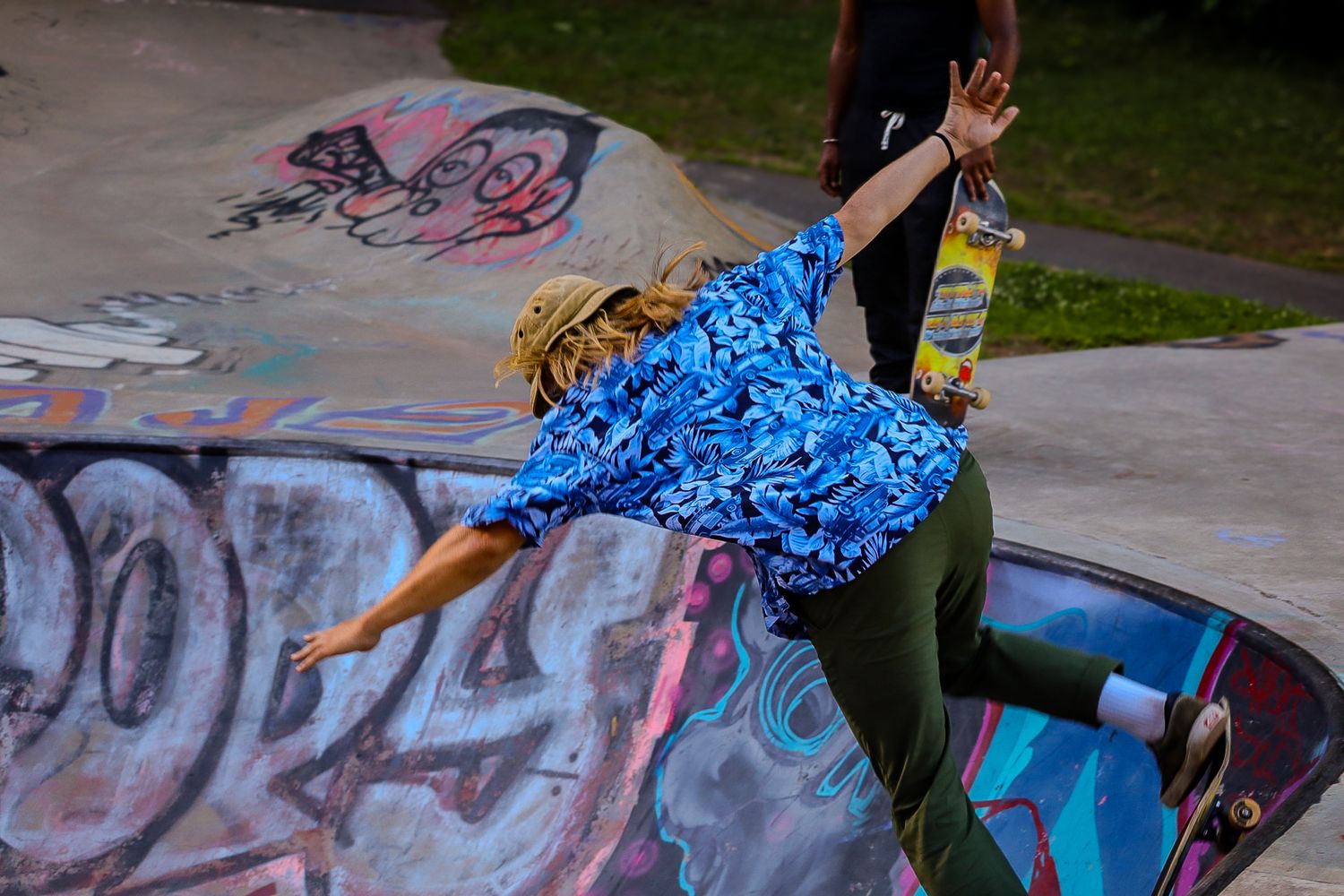 Skateboarder performing a trick in blue patterned shirt at graffiti-covered skate park.