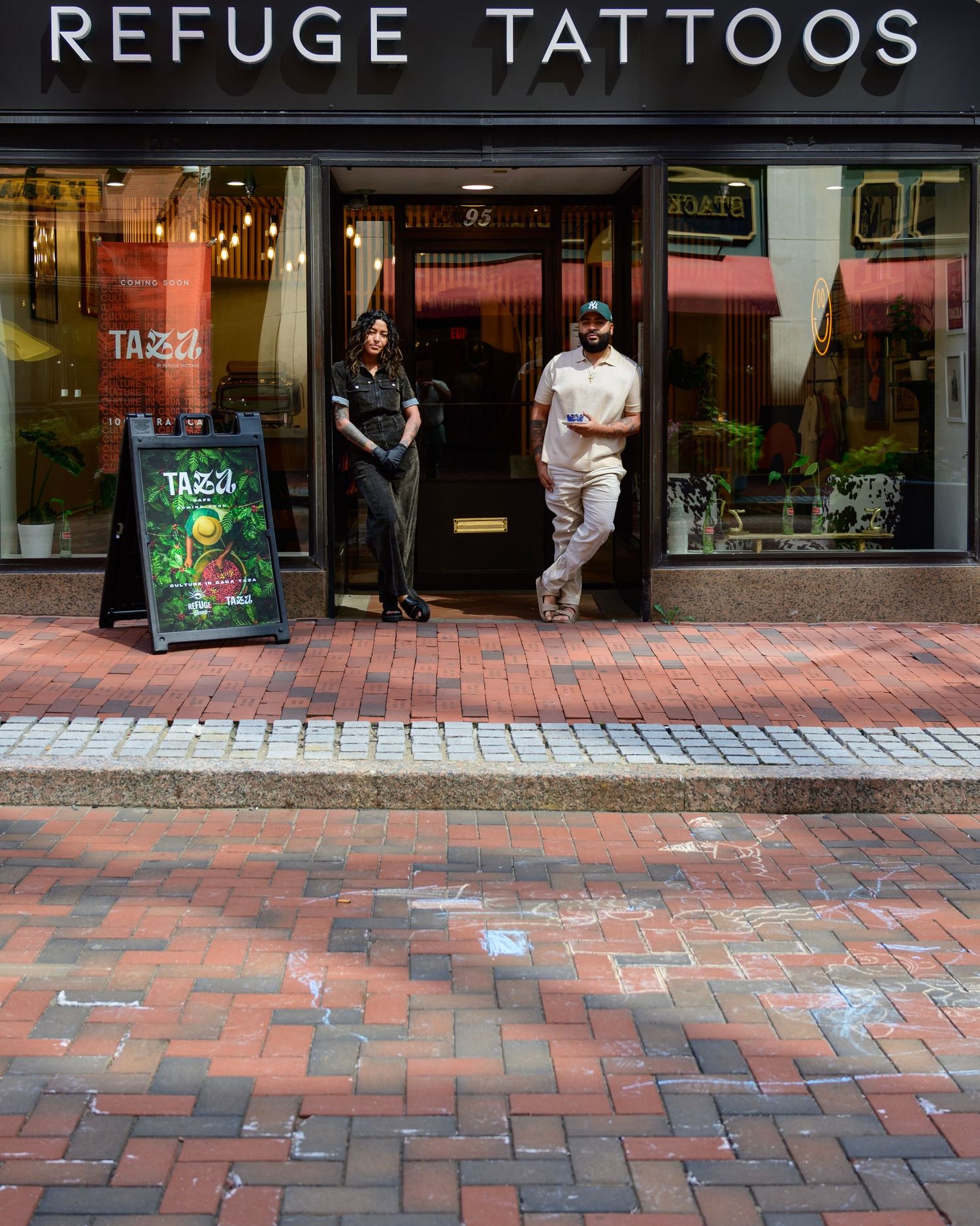 Two people exit Refuge Tattoos shop with a Tazza sandwich board sign on a red brick sidewalk.