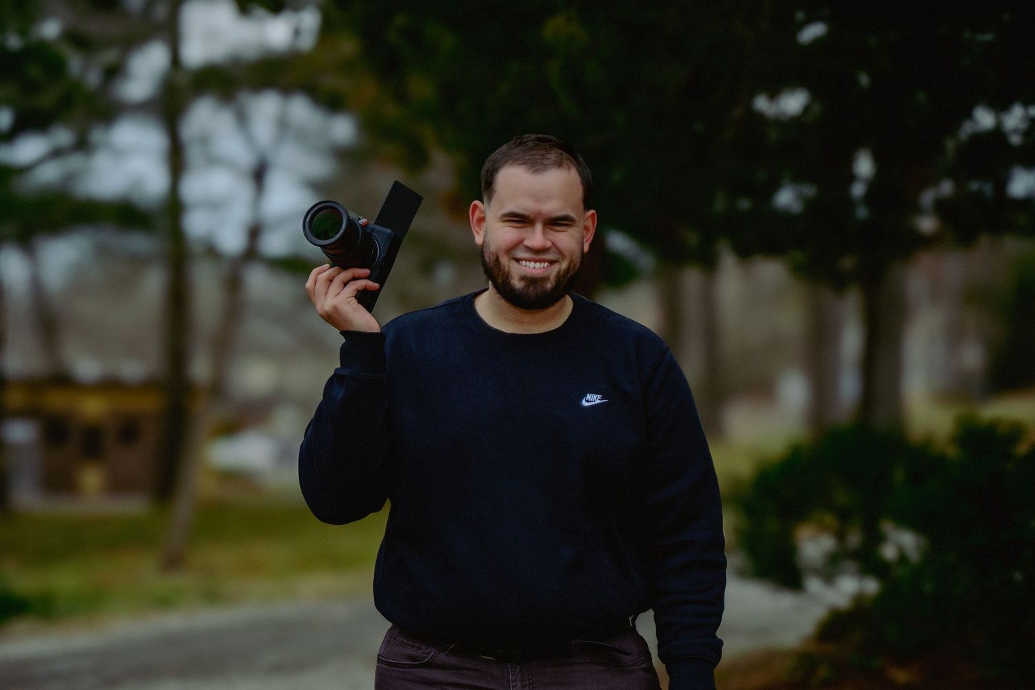A person in a black sweater stands outdoors against a blurred natural background.