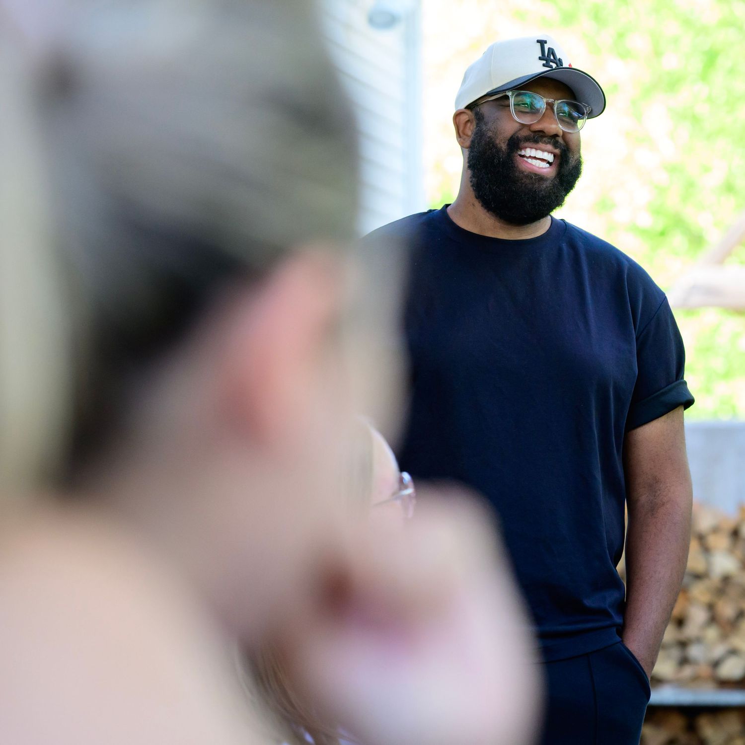 Smiling bearded Black man wearing LA cap and navy t-shirt laughing outdoors in bright sunlight.