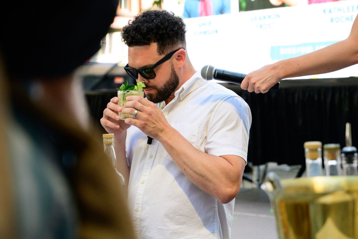 Man in white shirt smelling a green cocktail drink at an outdoor bar event with microphone nearby.
