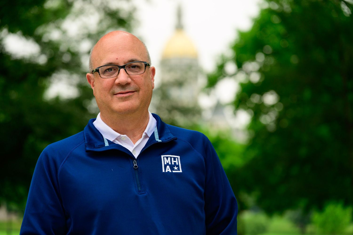 Man in blue jacket smiling outdoors with golden dome building visible among green trees in background.