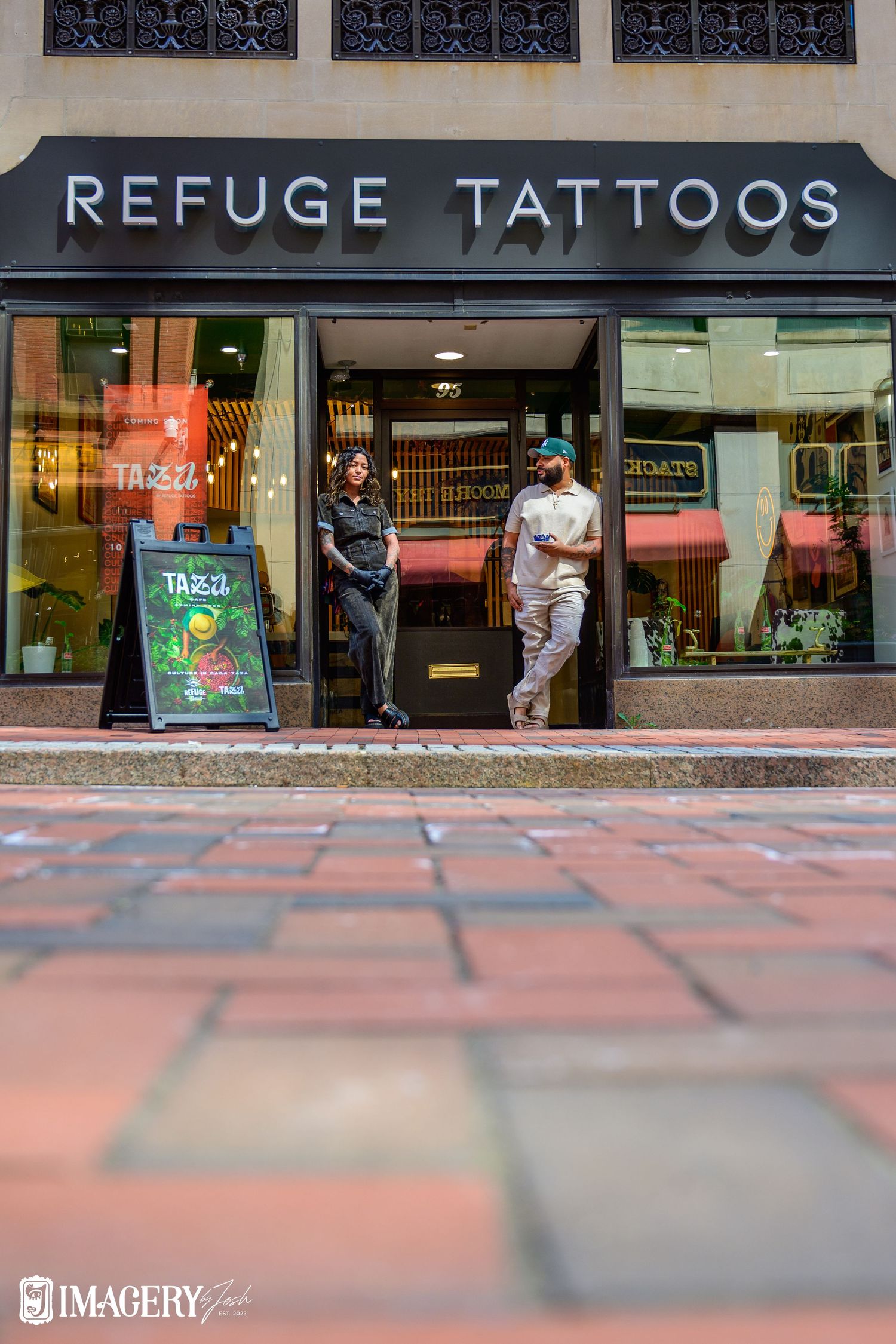 Two people stand outside Refuge Tattoos shop entrance on a brick sidewalk in an urban setting.