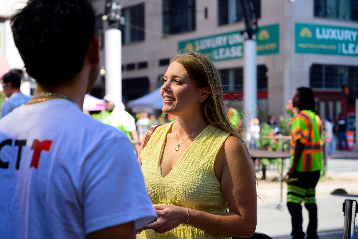 A smiling blonde woman in a yellow dress converses outdoors at a sunny urban street event.