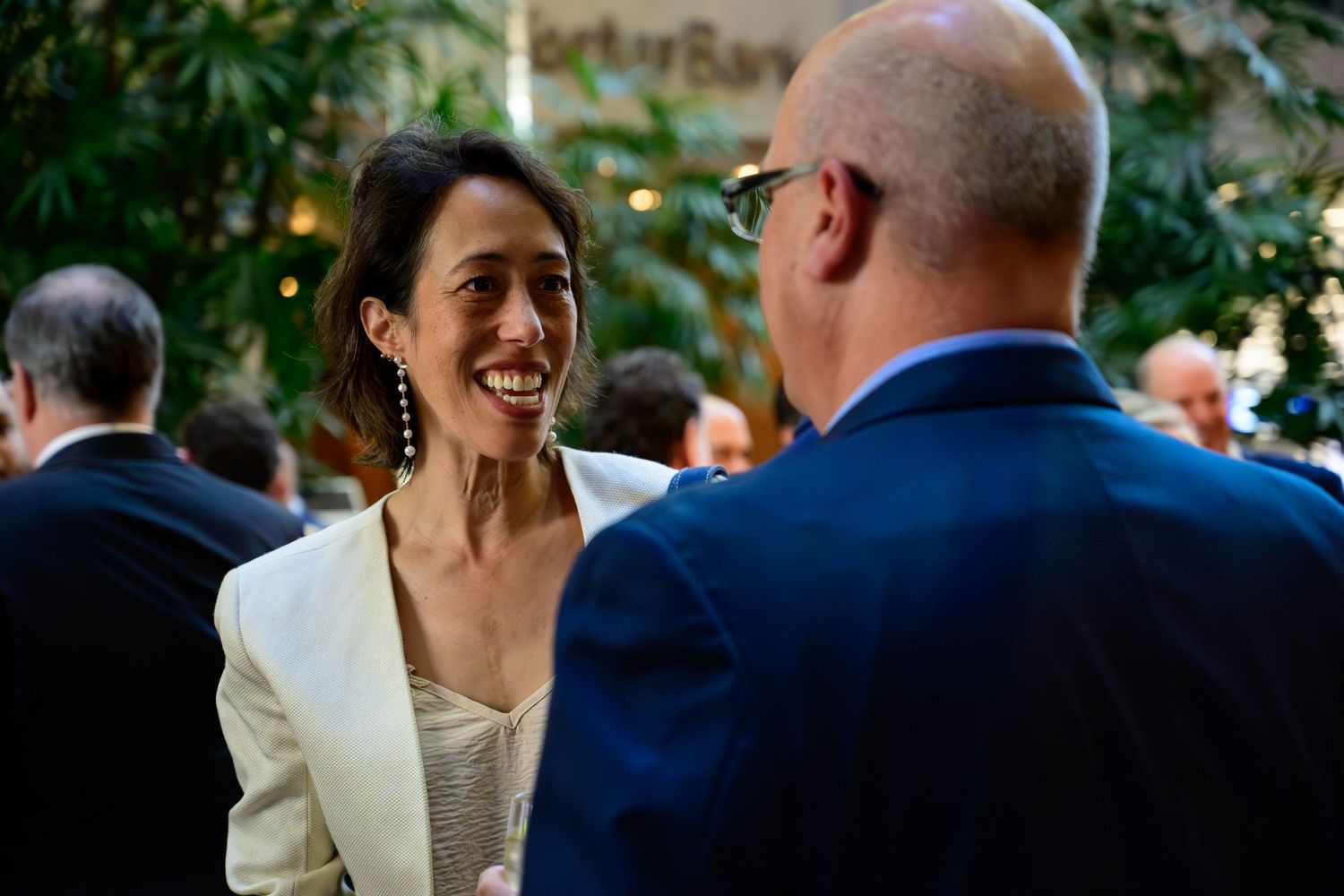 Woman in white blazer smiling and conversing with a man in a blue suit at an elegant indoor networking event.