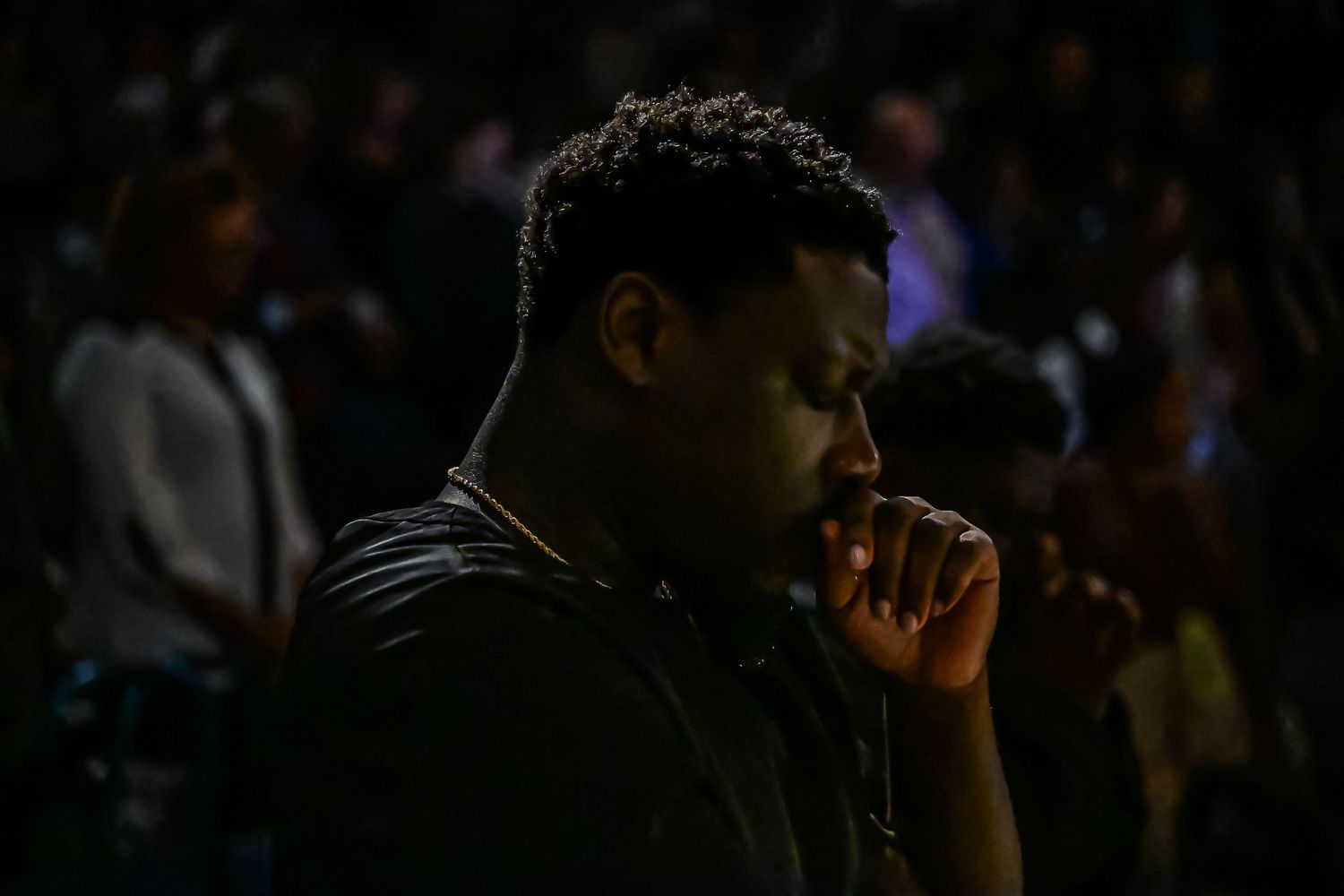 A silhouetted figure in deep thought stands against a dark background with a crowd visible behind them.